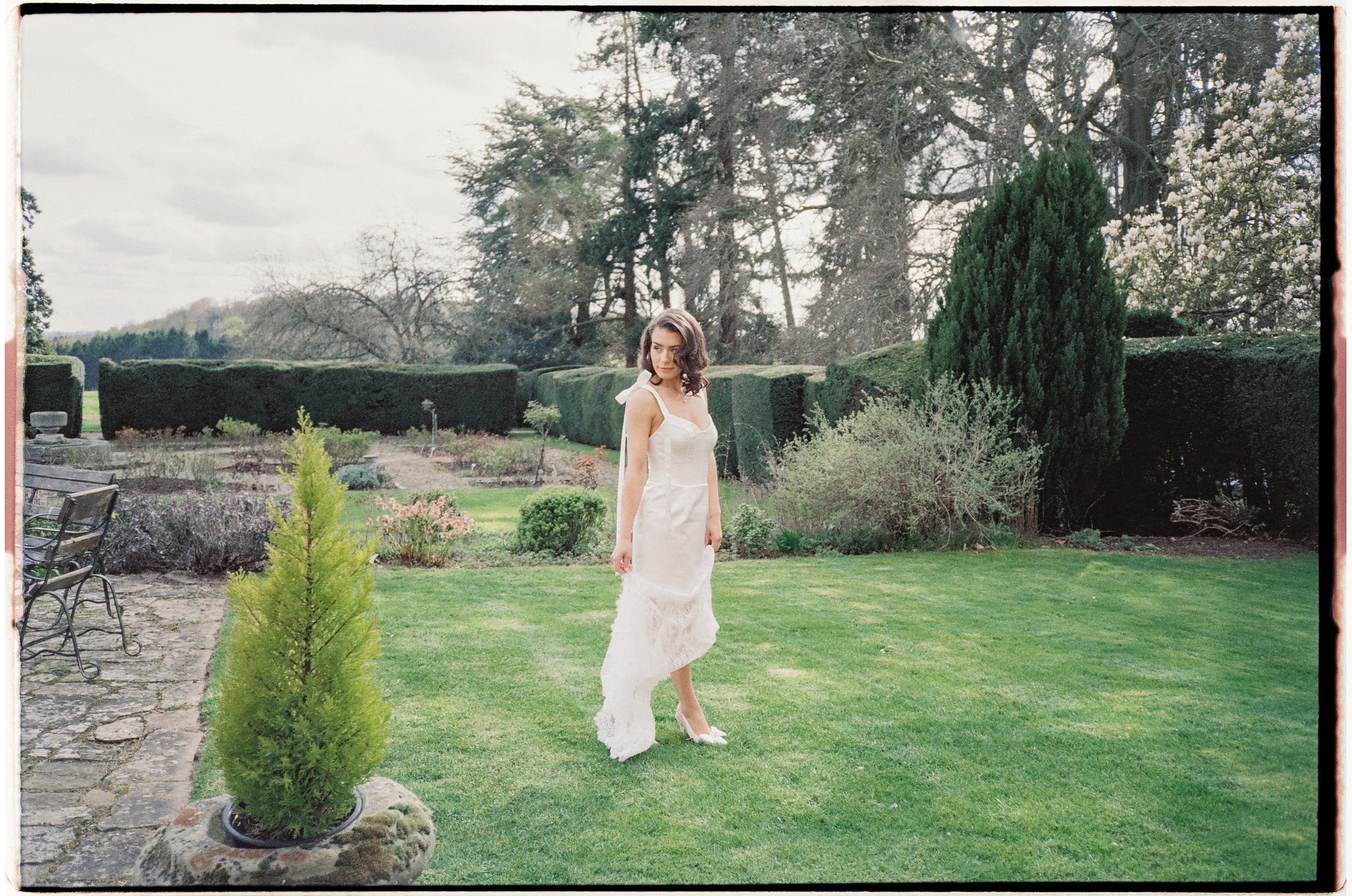 A woman in a white dress standing on a well-maintained lawn in a garden with various bushes, trees, and flowering plants, under a cloudy sky.