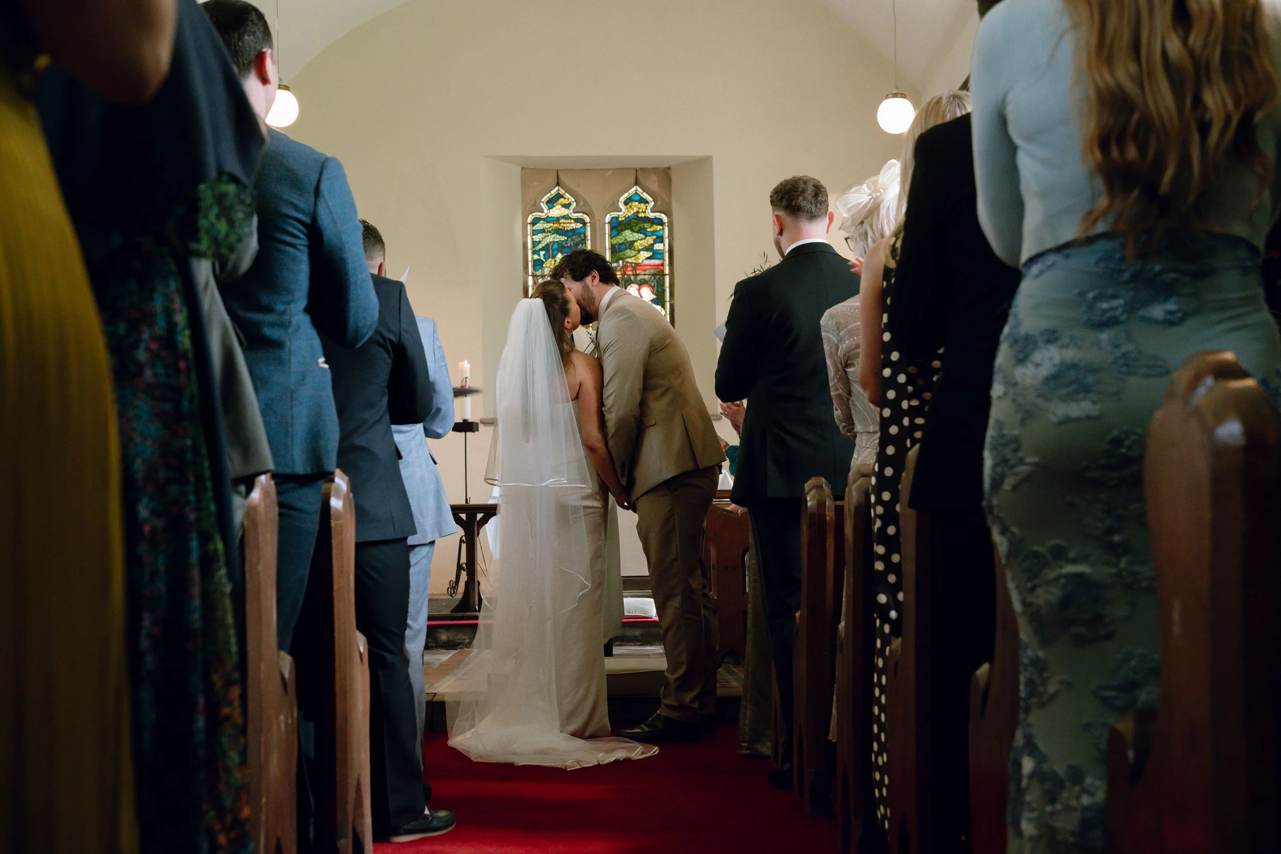 A bride and groom share a kiss during their wedding ceremony inside a church, surrounded by seated guests on either side.