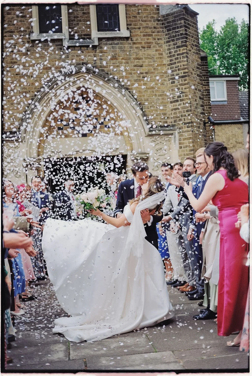 A wedding celebration outside a church with confetti falling as the bride and groom kiss, surrounded by friends and family including a woman in a pink dress clapping and smiling.