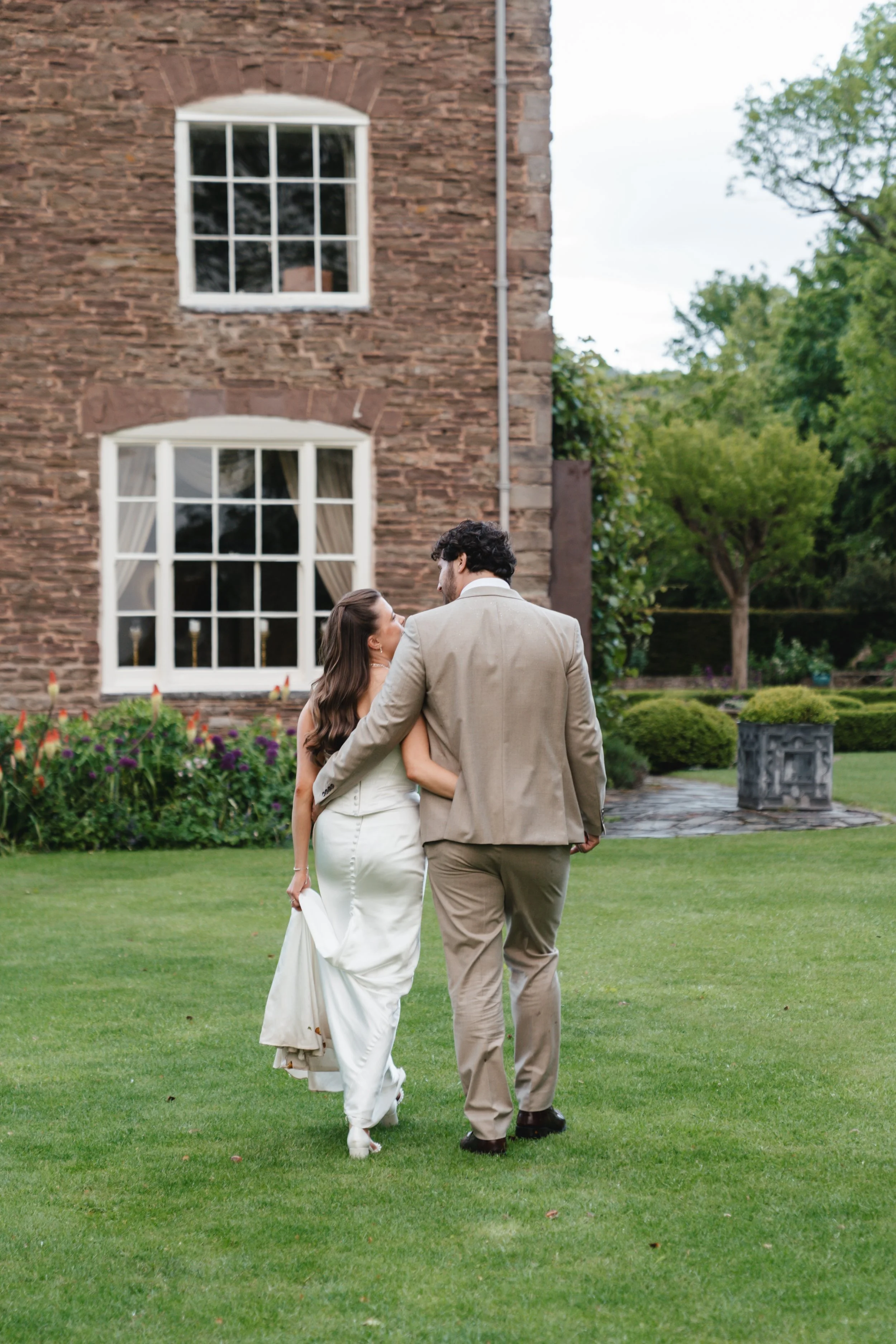 A bride and groom walking on a lush green lawn, holding hands, with a large brick house and blooming garden in the background.