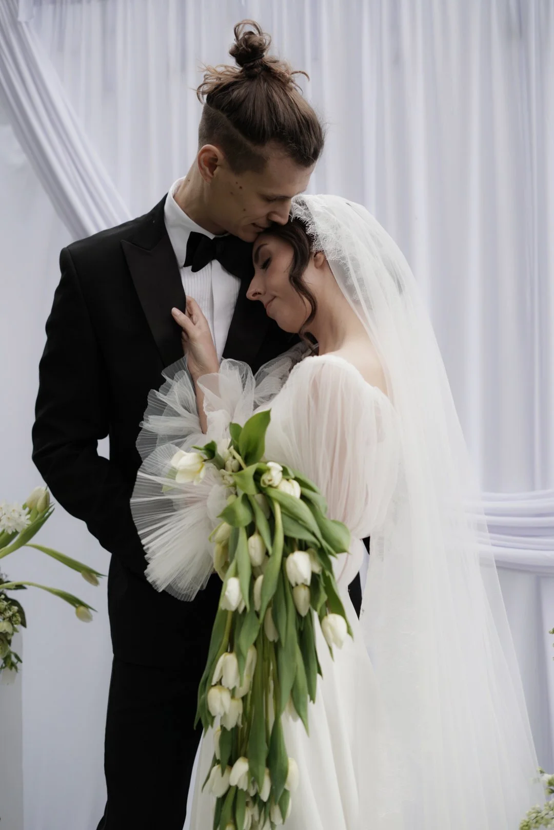 A bride and groom share an emotional moment, with the groom gently kissing the bride's forehead. The bride wears a white wedding gown and veil, holding a large bouquet of white tulips, while the groom is dressed in a black tuxedo with a bow tie, against a white draped background.