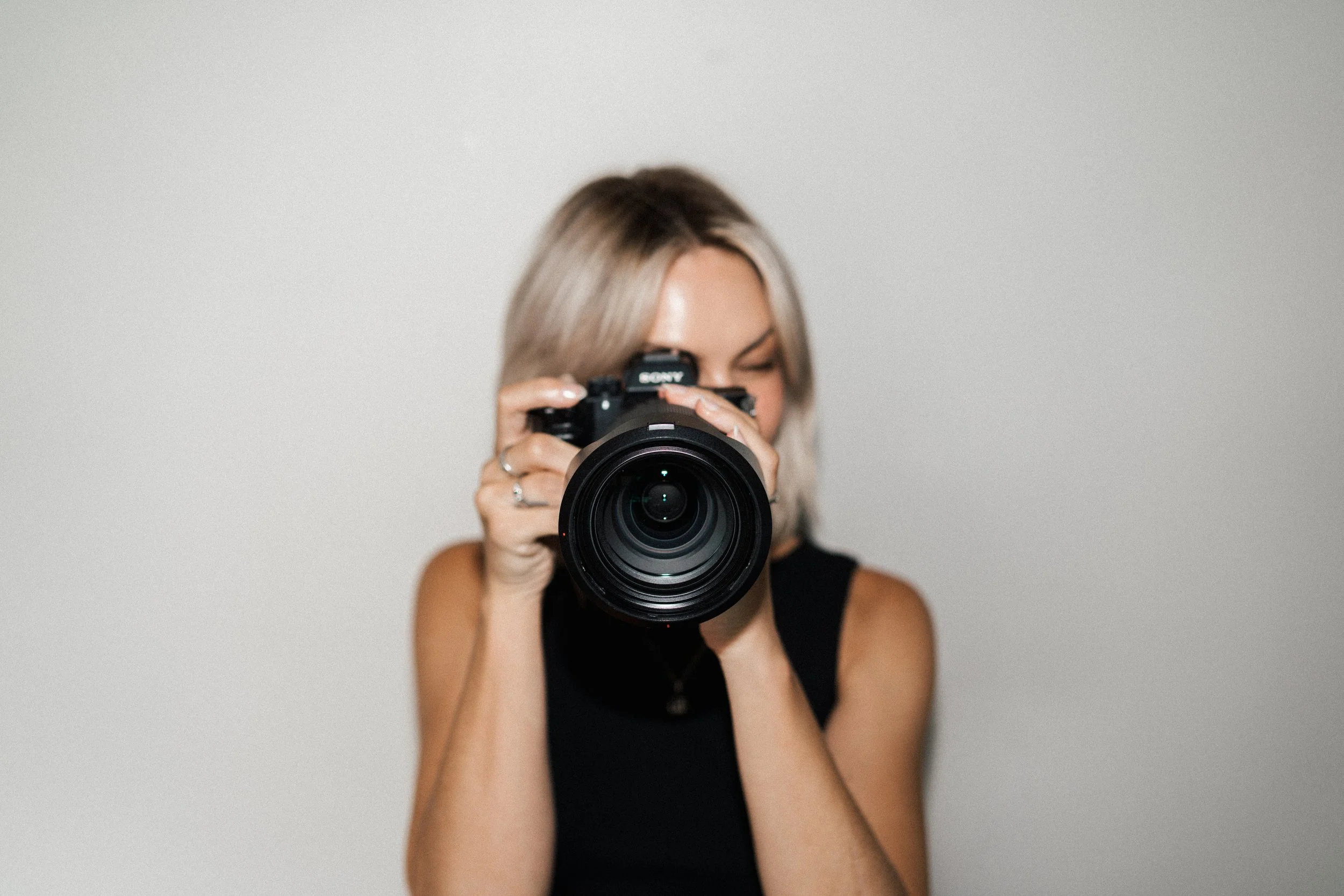 A woman with blonde hair takes a photo using a professional camera against a plain light gray background.