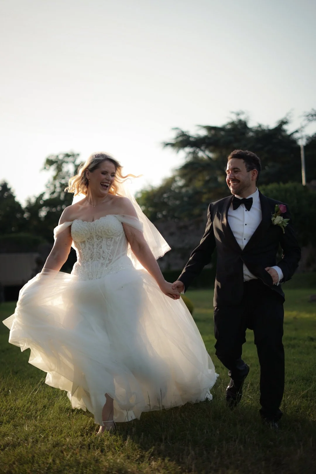 A bride and groom holding hands and smiling outdoors on a sunny day, with trees and grassy area in the background.