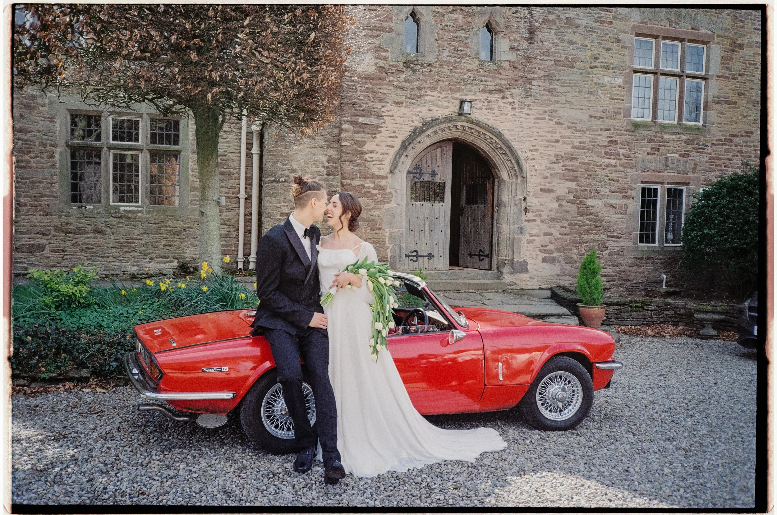 A newlywed couple dressed in wedding attire standing beside a red vintage convertible car in front of a stone building with multiple windows and a large arched door. The bride is holding a bouquet of white flowers, and they are sharing a joyful momen