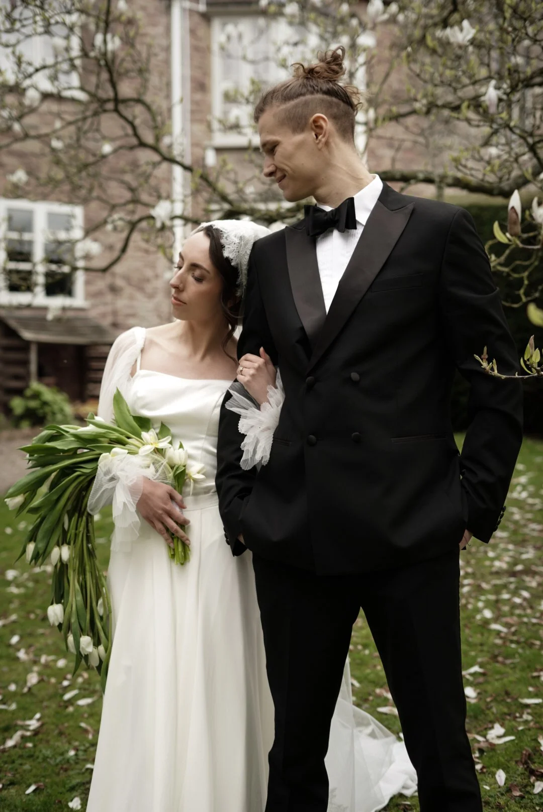 A bride holding a bouquet of white tulips and a groom dressed in a black tuxedo with a bow tie, standing outdoors near blossoming trees with white flowers.