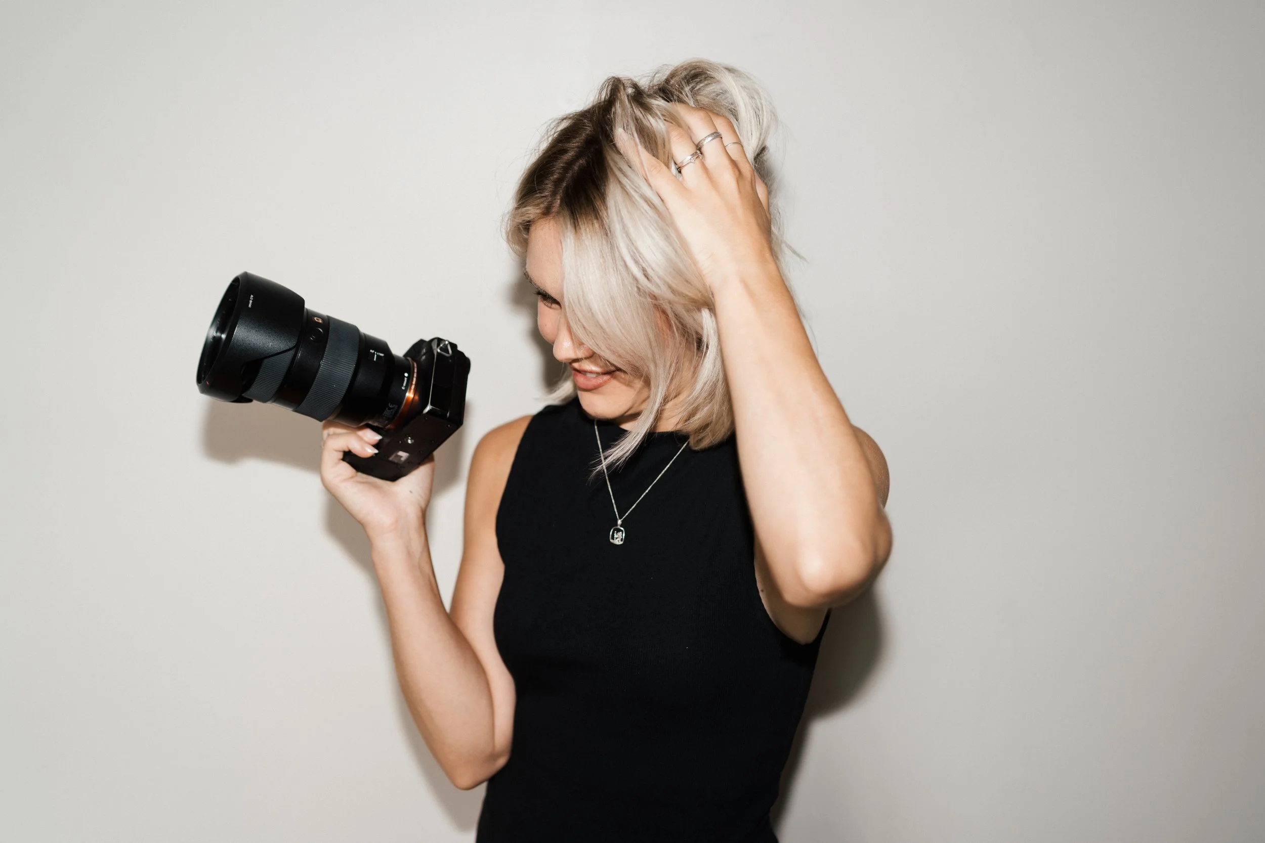 Woman with blonde hair, wearing a black sleeveless top, holding a camera in her right hand, smiling and touching her hair with her left hand, standing against a plain white wall.