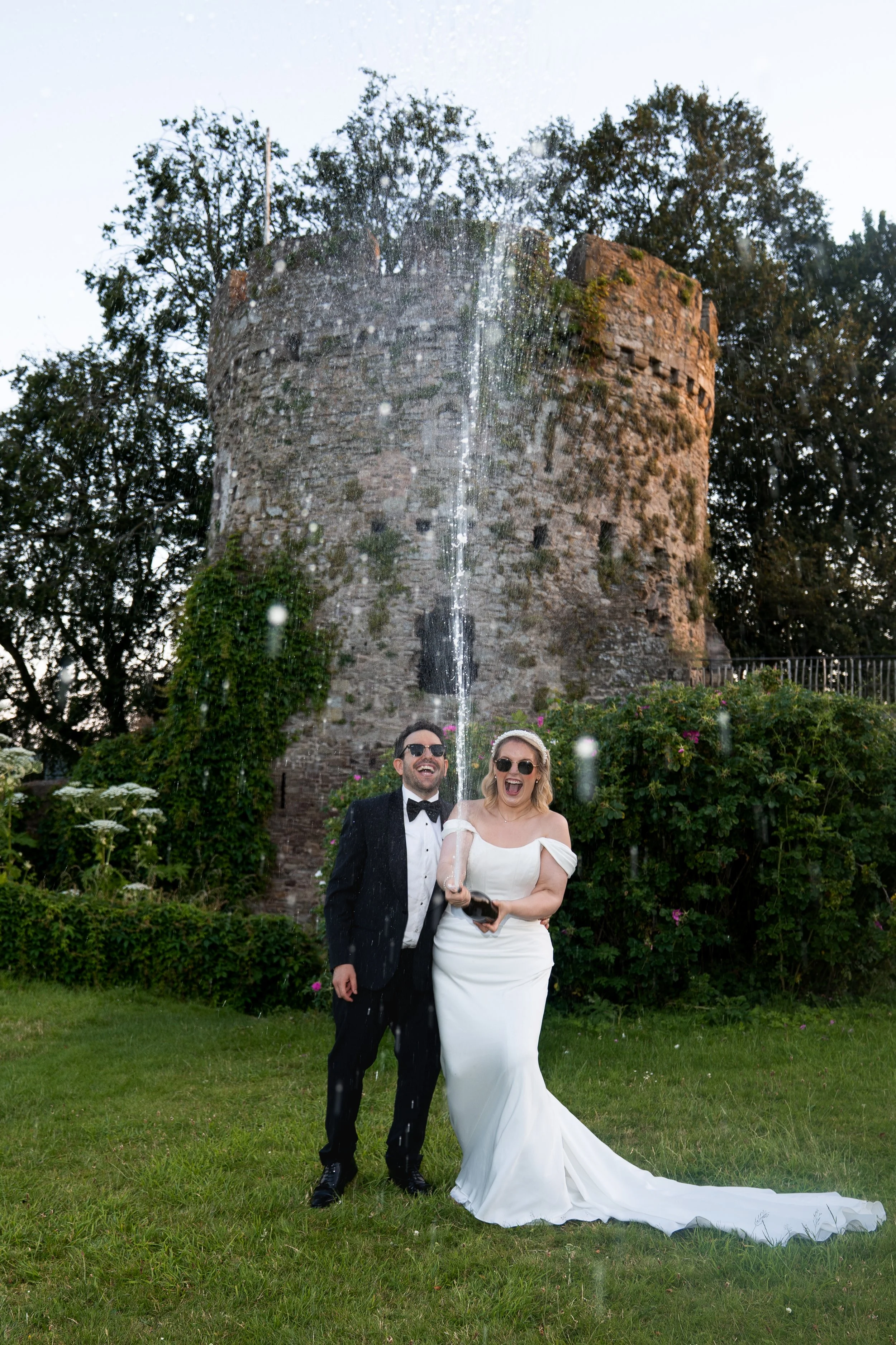 A bride and groom celebrating under a water fountain with an old stone tower and lush greenery in the background.