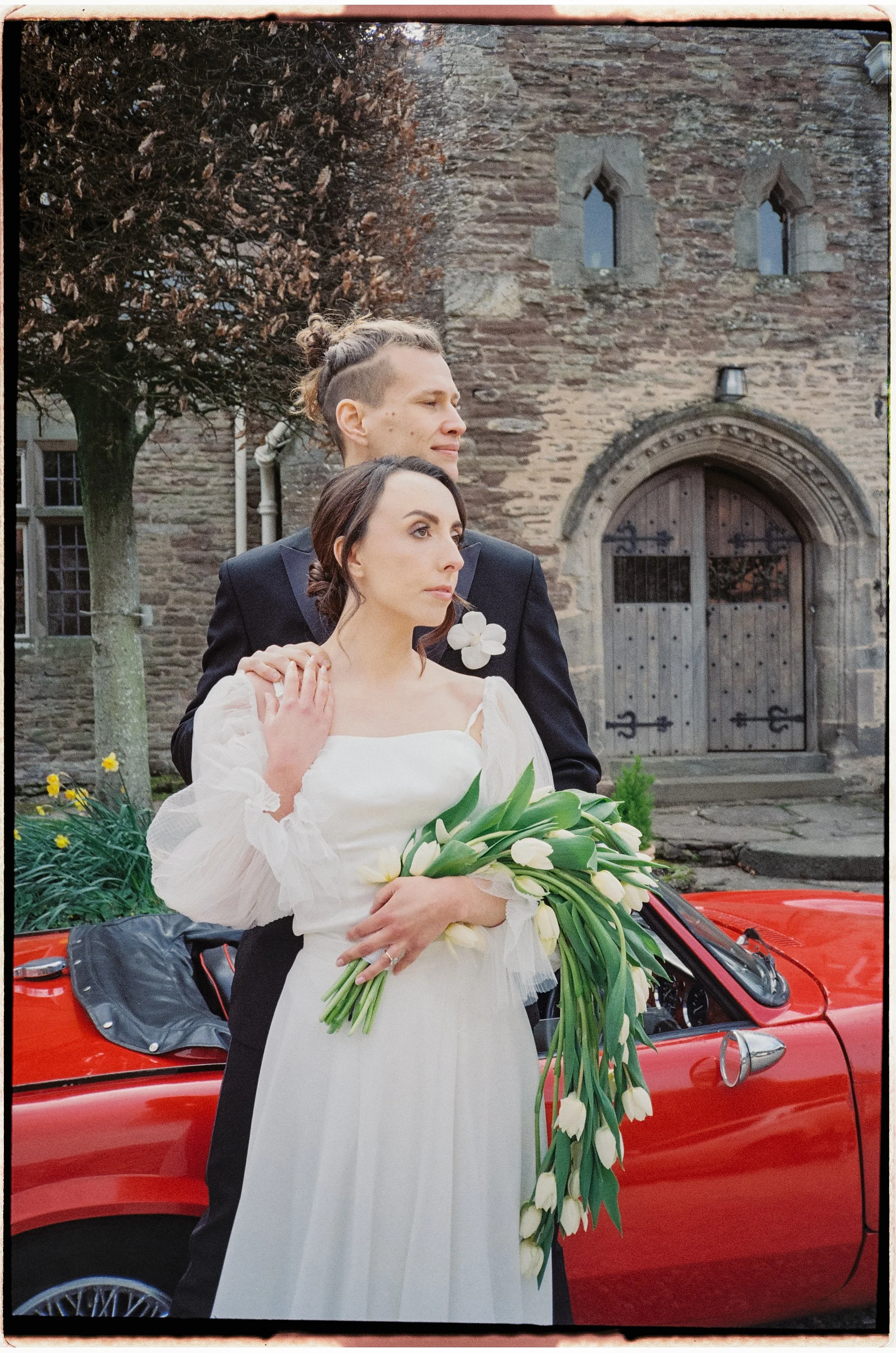 A man and woman in wedding attire stand outdoors in front of a red convertible car, holding a bouquet of white tulips, with an old stone building with arched wooden doors and small windows in the background.
