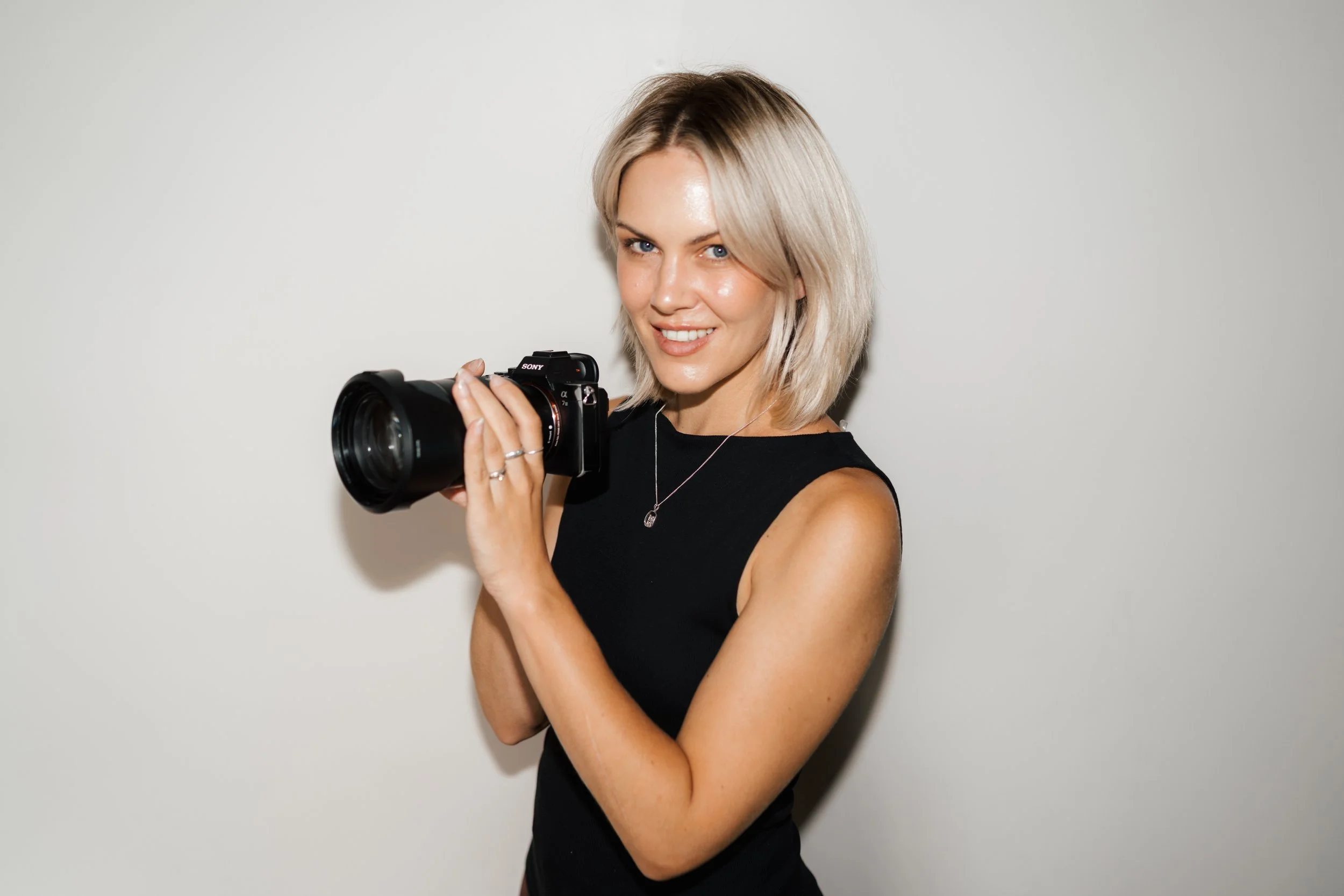 A young woman with shoulder-length blonde hair, wearing a black sleeveless top, holding a professional camera with both hands, smiling at the camera, standing against a plain light gray background.