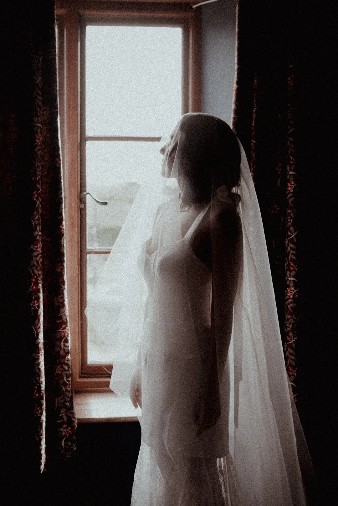 A woman wearing a wedding dress and veil is standing inside by a window, looking outside.
