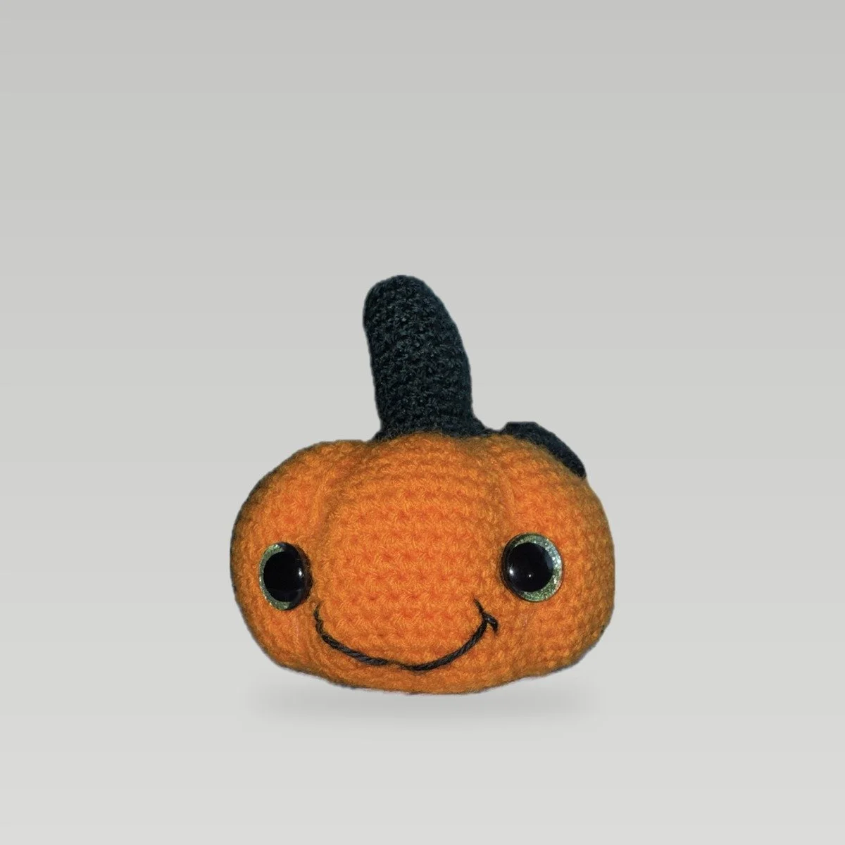 Handmade crochet pumpkin in orange with a dark green stem and leaf, green-glitter safety eyes, and a stitched smile