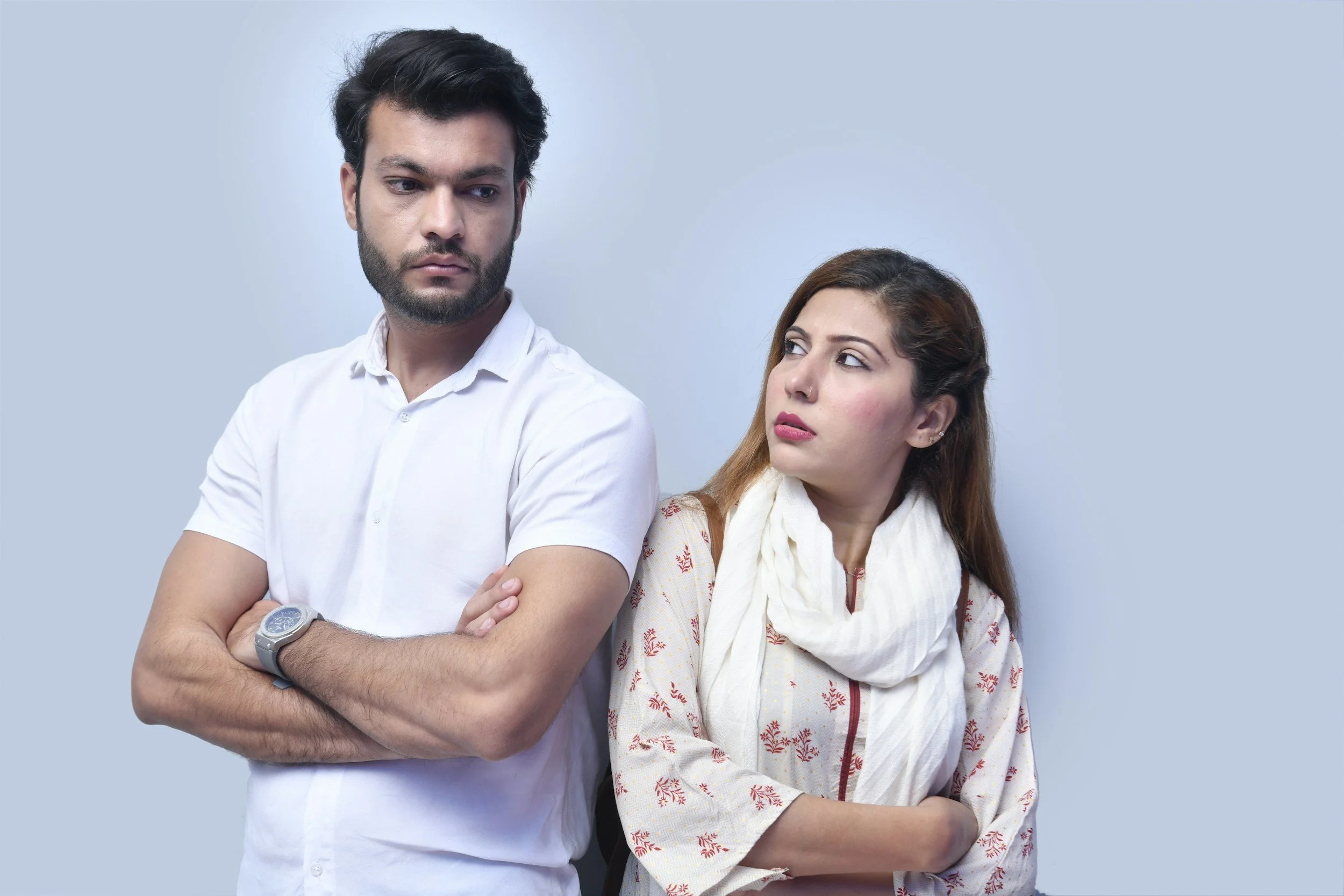 A young man with dark hair, beard, and a white shirt, standing with arms crossed, looking to the side. A young woman with brown hair, wearing a white scarf and a light-colored shirt with a red pattern, stands beside him, looking up at him with a concerned expression. They are in front of a plain light blue background.