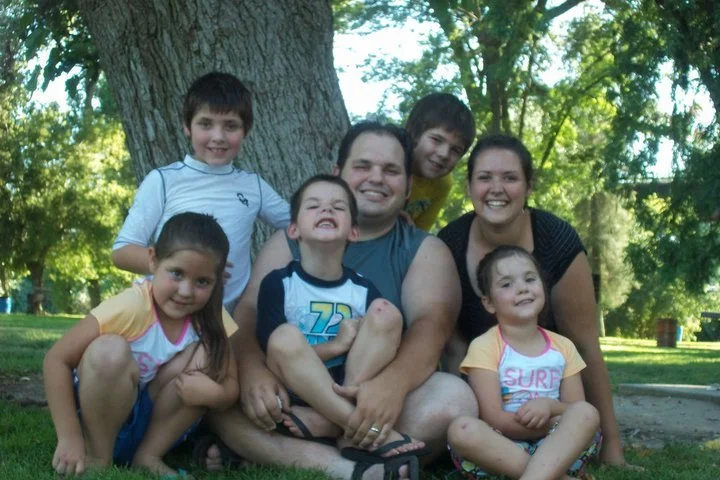 Family of six, including two adults and four children, sitting and standing in a park with green trees in the background.