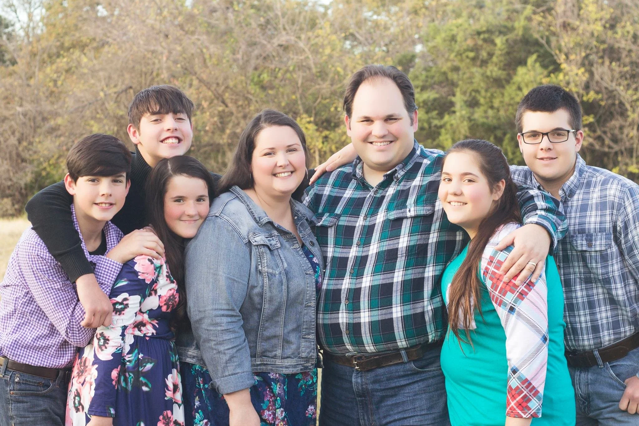 A large family group outdoors, smiling, with trees in the background. The group includes adults and children, all looking at the camera.