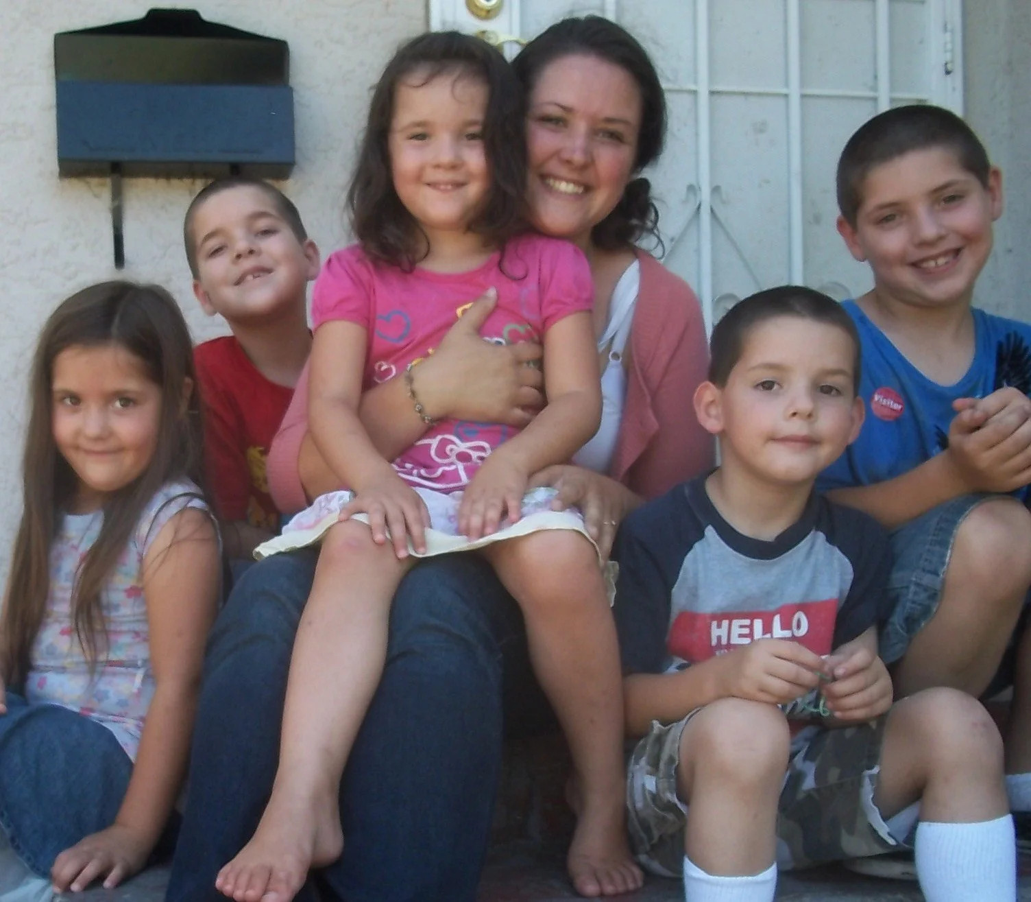 A group of six children and one woman sitting and posing for a photo outdoors against a wall and gate, smiling at the camera.