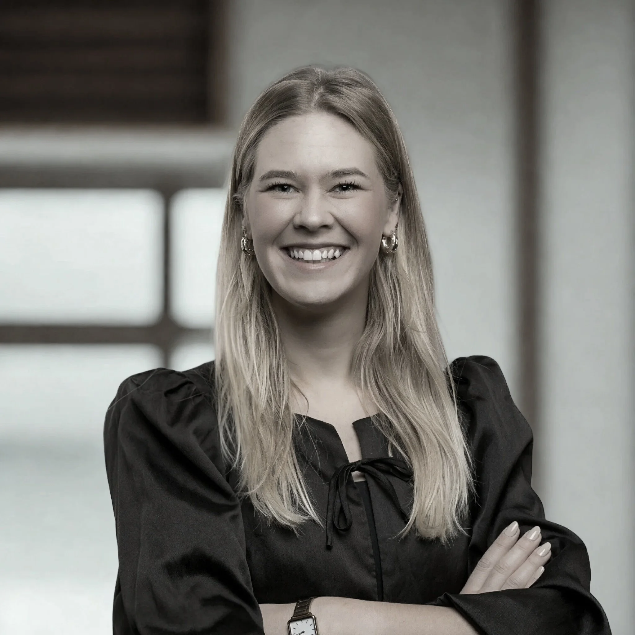 Close-up van een glimlachende vrouw met lang blond haar, zwart jasje, horloge en oorbellen, met haar armen over elkaar, op een witte achtergrond.