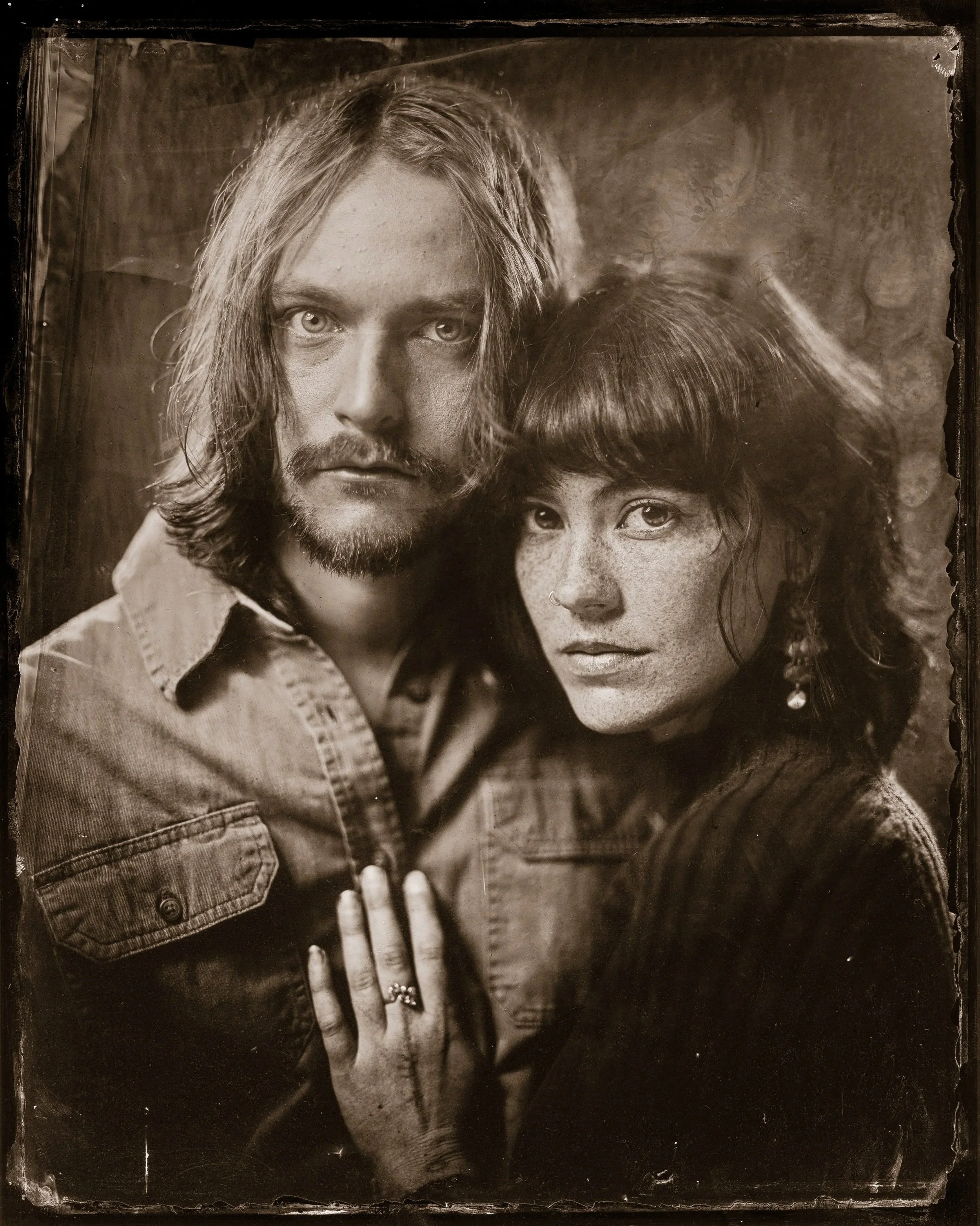 A black and white vintage photograph of a man and a woman close together, with the man on the left and the woman on the right. The man has long hair, a beard, and is wearing a denim jacket. The woman has short curly hair, freckles, and is wearing a d