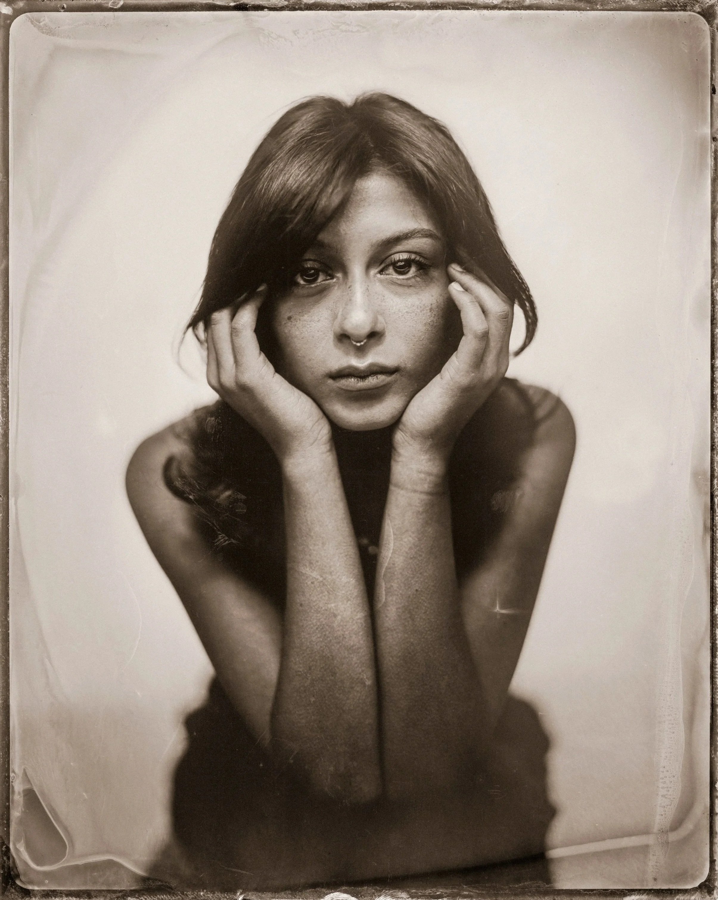Black and white photo of a woman with dark hair holding her face with her hands, staring directly at the camera.