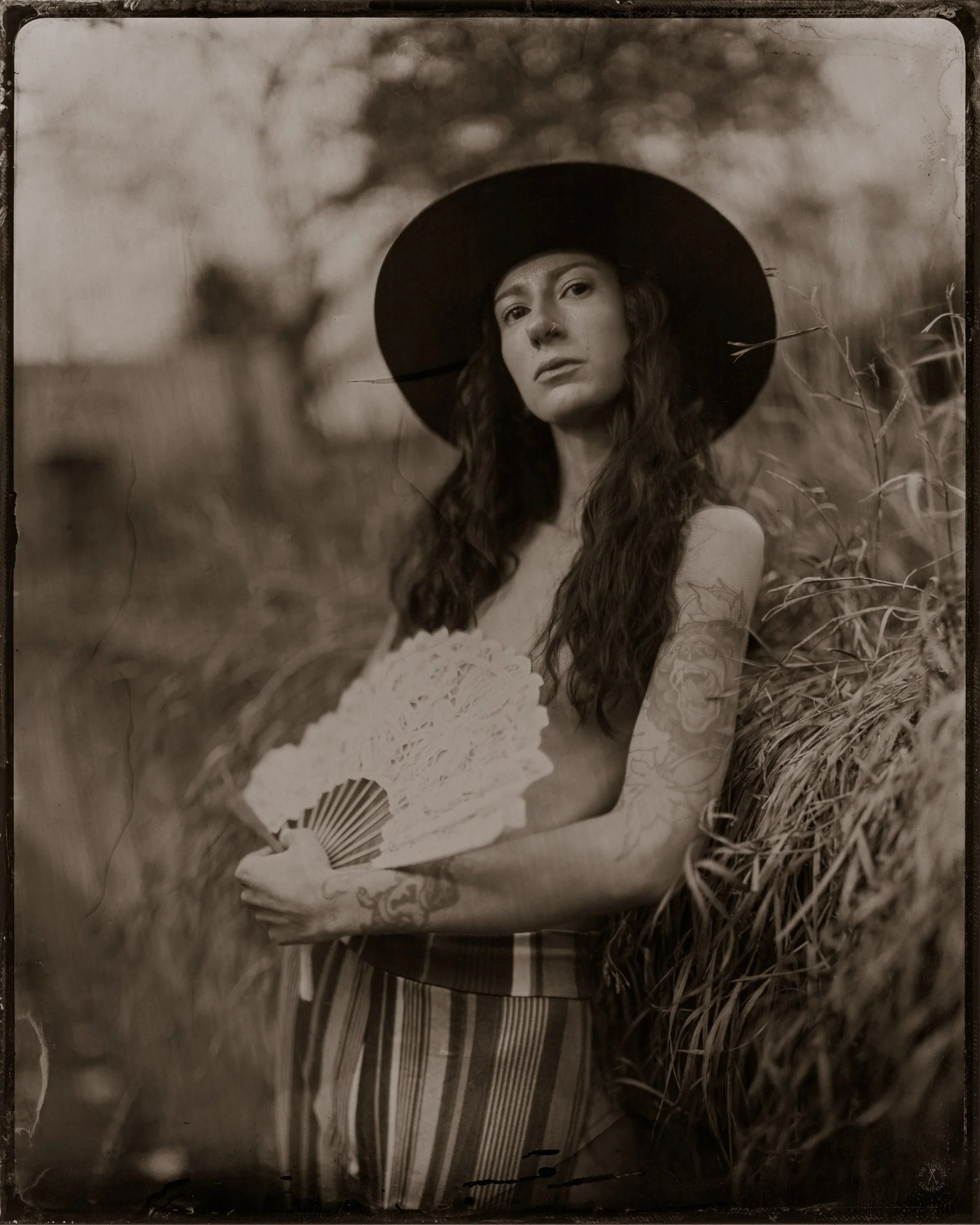 Black and white photograph of a woman with long wavy hair wearing a wide-brimmed hat and holding a lace fan, standing outdoors among tall grass or wheat. The image has a vintage or artistic style.