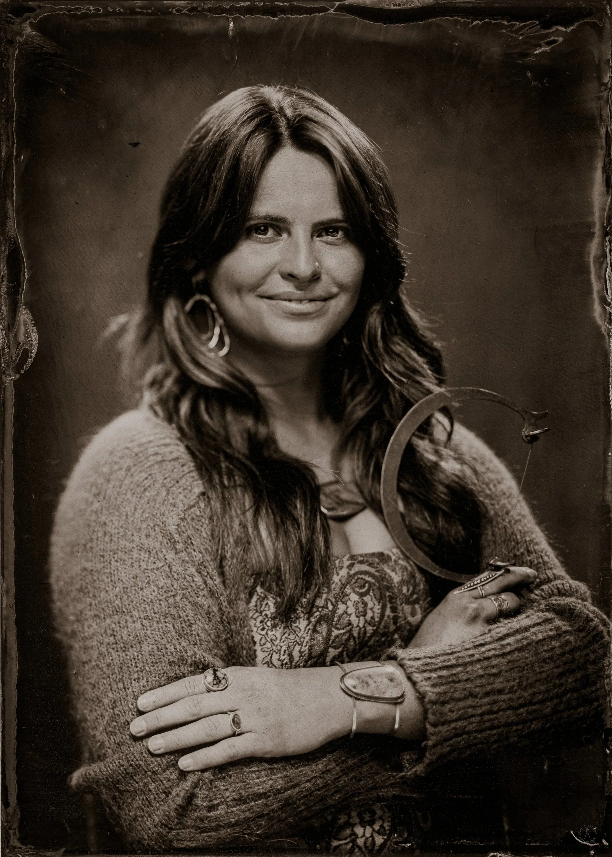 Black and white photograph of a woman with long wavy hair, wearing earrings, a patterned top, and a sweater. She is smiling slightly, with her arms crossed, holding a pair of glasses and wearing rings. The image has a vintage, slightly worn appearanc