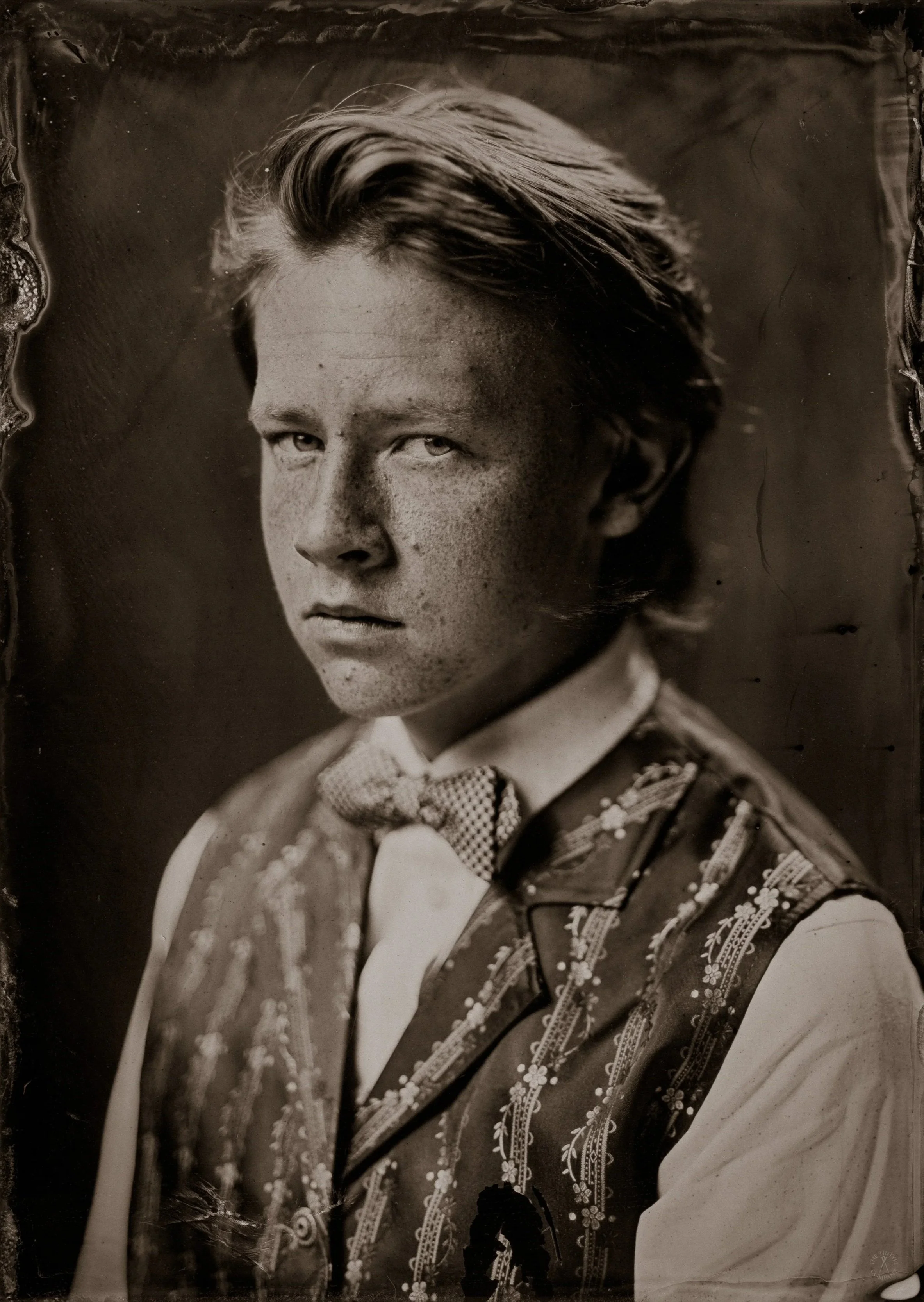 Black and white portrait of a young man with styled hair, dressed in ornate traditional clothing with embroidered details and a bow tie.