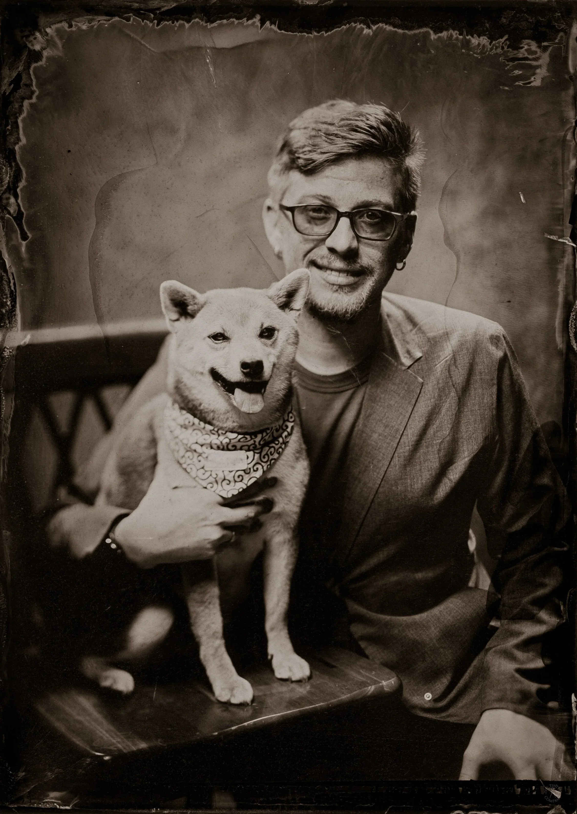 A black and white photograph of a smiling man with glasses, wearing a jacket, holding a dog with a bandana around its neck. The dog is sitting on a flat surface, and the man has his arm around it.