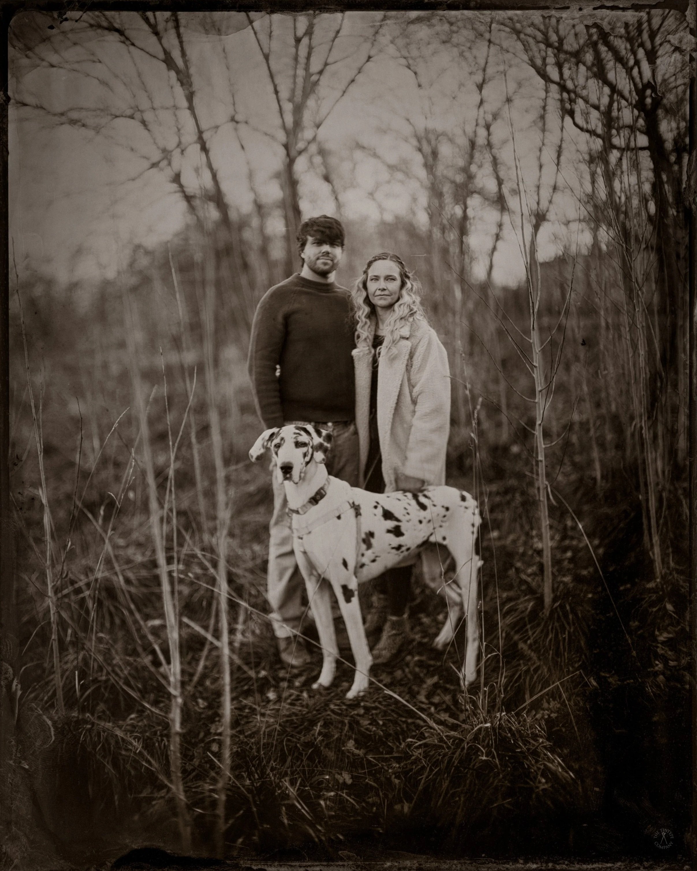 Black and white photo of a young man with dark hair and beard, a woman with wavy blonde hair, and a Dalmatian dog, standing outdoors in a forest with leafless trees and tall grass.
