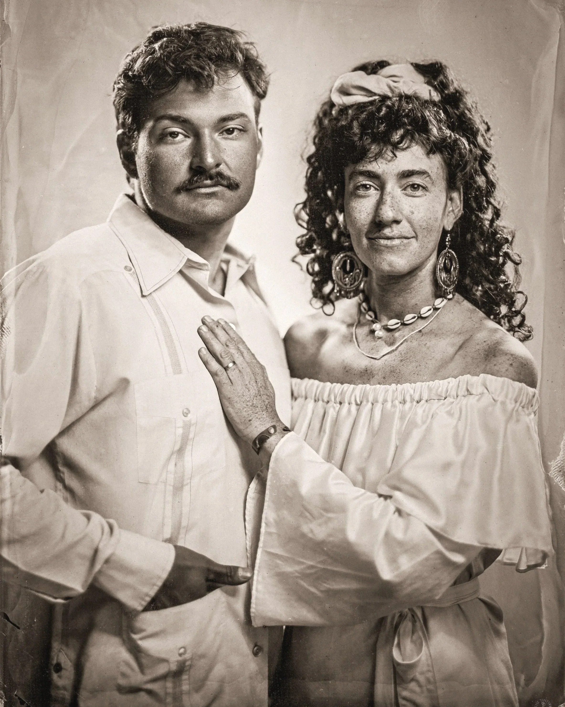 A black-and-white photograph of a man with dark hair, mustache, and light skin, and a woman with long curly hair, earrings, and a necklace. Both are dressed in vintage clothing, with the woman in an off-shoulder top, striking a confident pose, with t