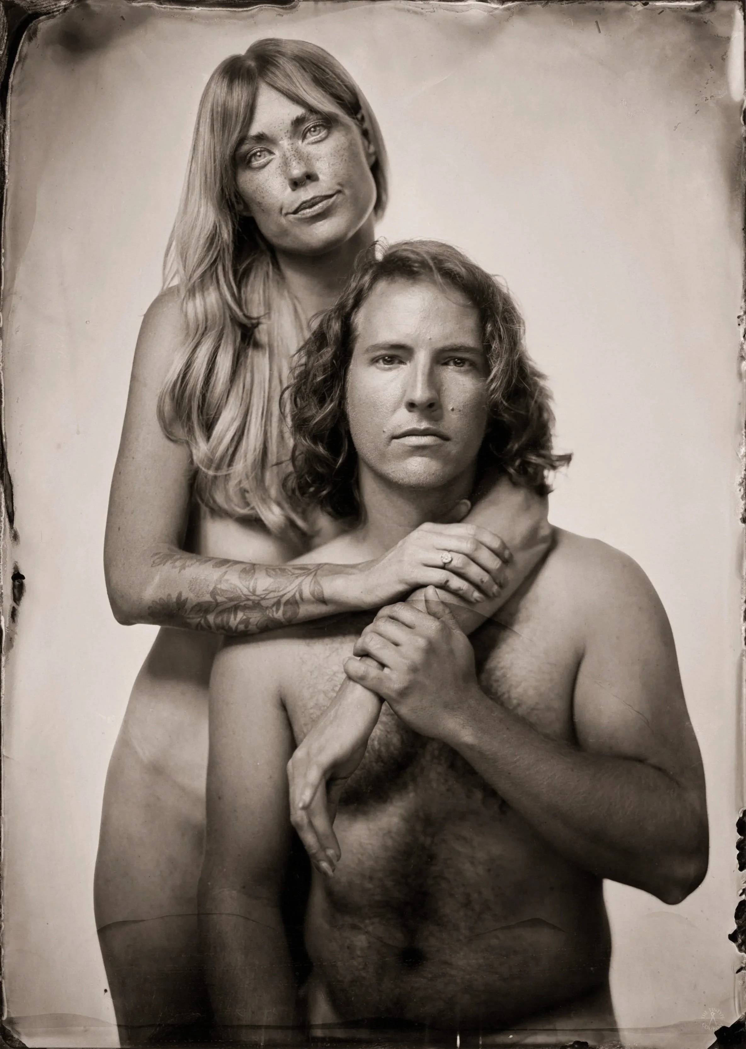 A black and white photograph of a woman standing behind a seated man, both nude. The woman has long, wavy hair and tattoos on her arm, and she is looking directly at the camera with a slight smile. The man has long, curly hair and a serious expressio