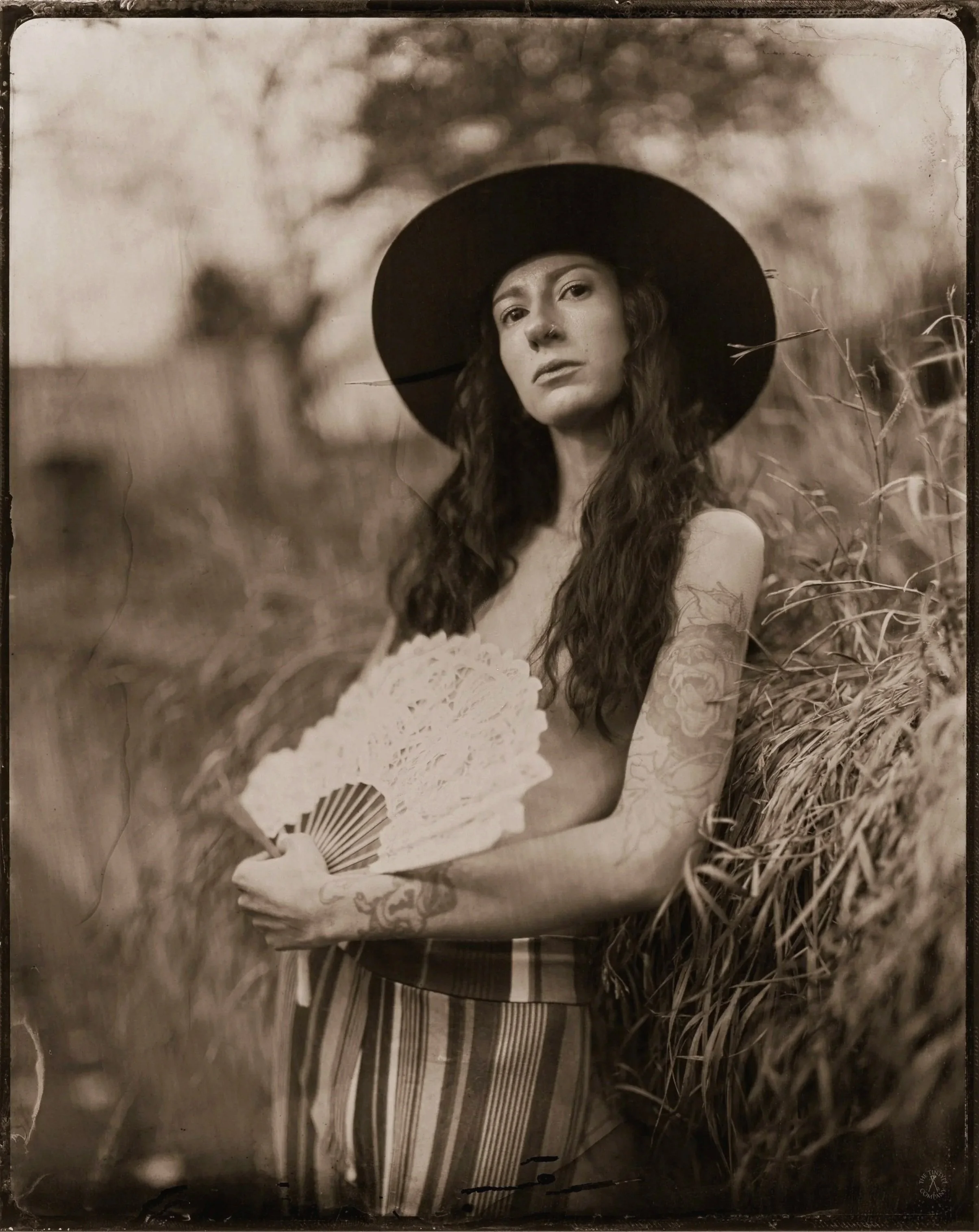 A woman with long hair, wearing a wide-brimmed hat, is holding a parasol in an outdoor natural setting with tall grass surrounding her.