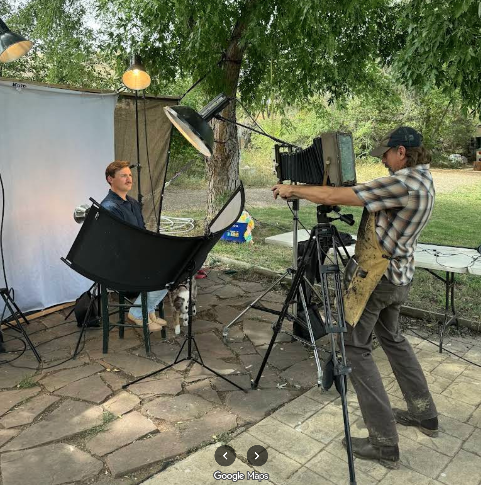 Two men setting up filming equipment outdoors under a large tree. One man is adjusting a camera on a tripod, while the other man is sitting on a chair next to a small dog with a black and white coat. There is filming lighting equipment and a white backdrop nearby.
