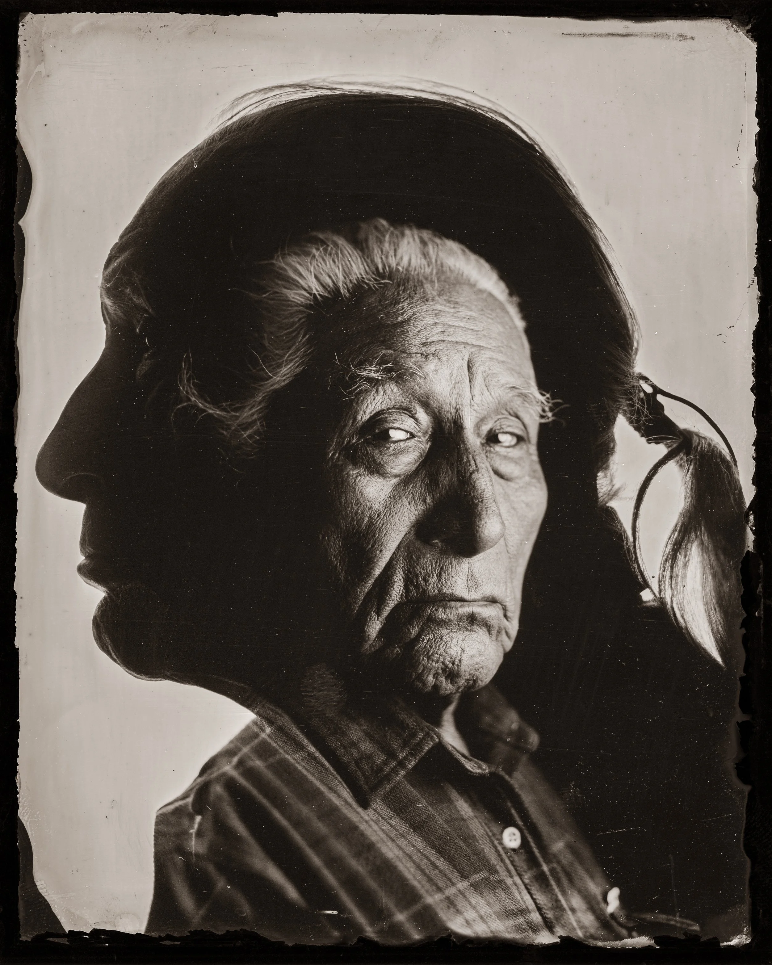 A black and white photograph of an elderly woman in traditional fashion, with her hair styled in a bun, dressed in a plaid shirt. She is looking to her left with a serious expression.