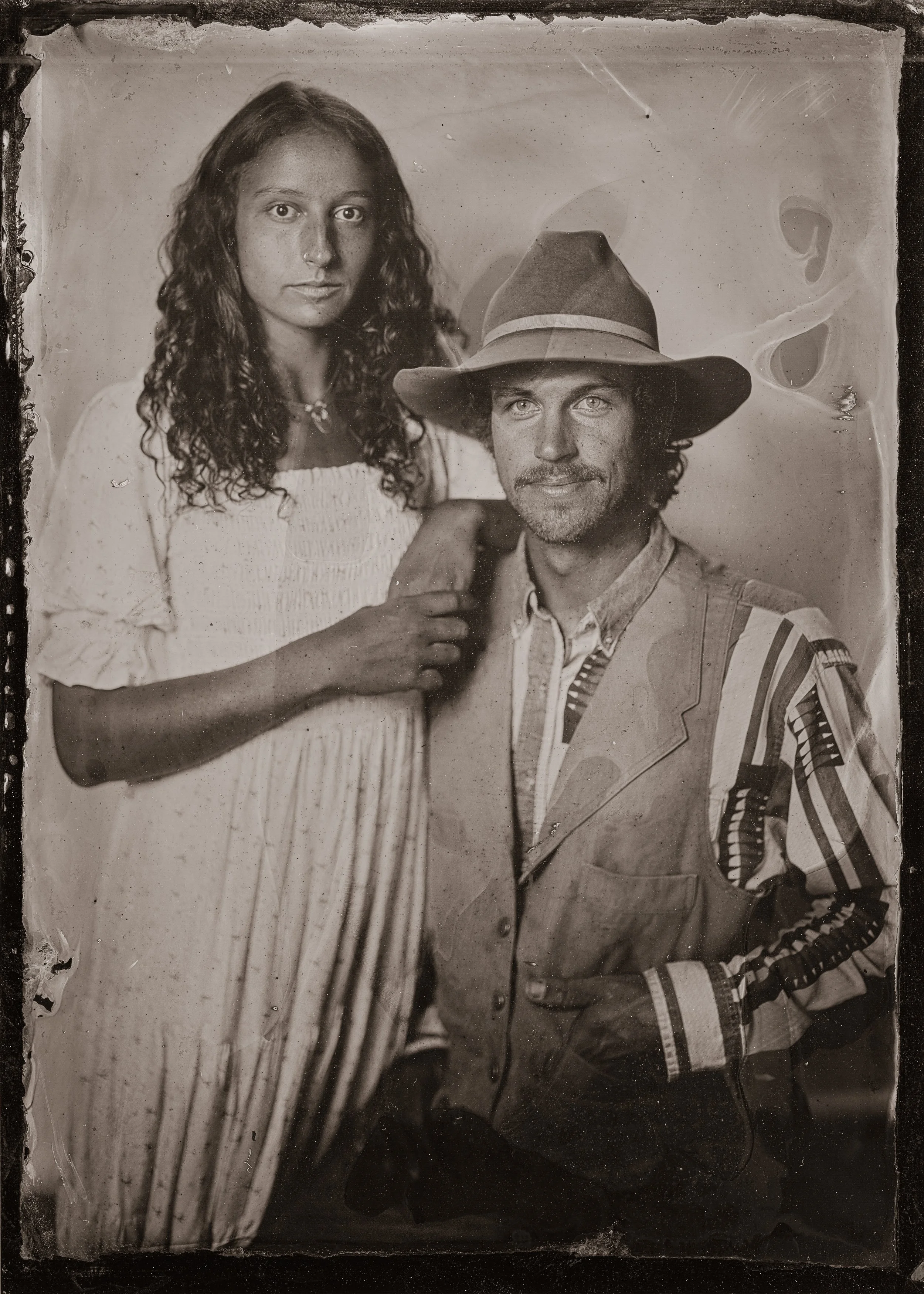 A vintage black and white photograph of a young woman with curly hair wearing a white dress and a large necklace, standing close to a man with a beard, wearing a wide-brimmed hat and a striped jacket with a patch on the shoulder.