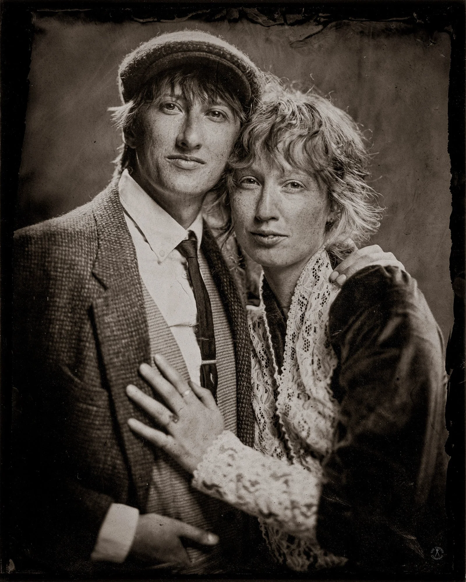Black and white portrait of a young man and woman standing close together, looking at camera. The man is wearing a hat, a patterned jacket, and a tie. The woman has curly hair and is wearing a lace blouse. They are outside, with a blurred background.