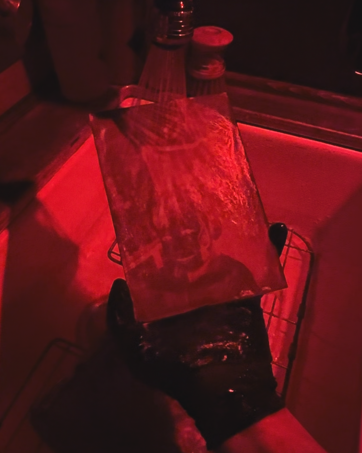 A reflective surface, like a mirror or metallic panel, on a cart in a dimly lit room illuminated by red light, with a person's legs visible at the bottom of the image.
