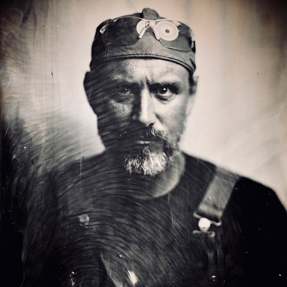 A black and white portrait of a man with a beard, dressed in a dark shirt and cap with glasses resting on top, looking directly at the camera.