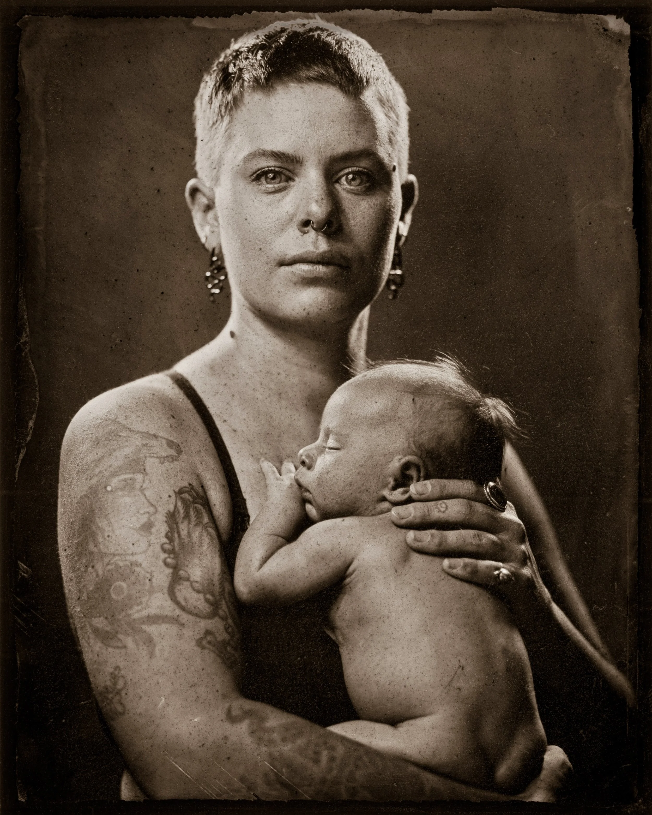 Black and white photo of a woman with short hair, wearing earrings and a septum piercing, holding a sleeping baby close to her chest.