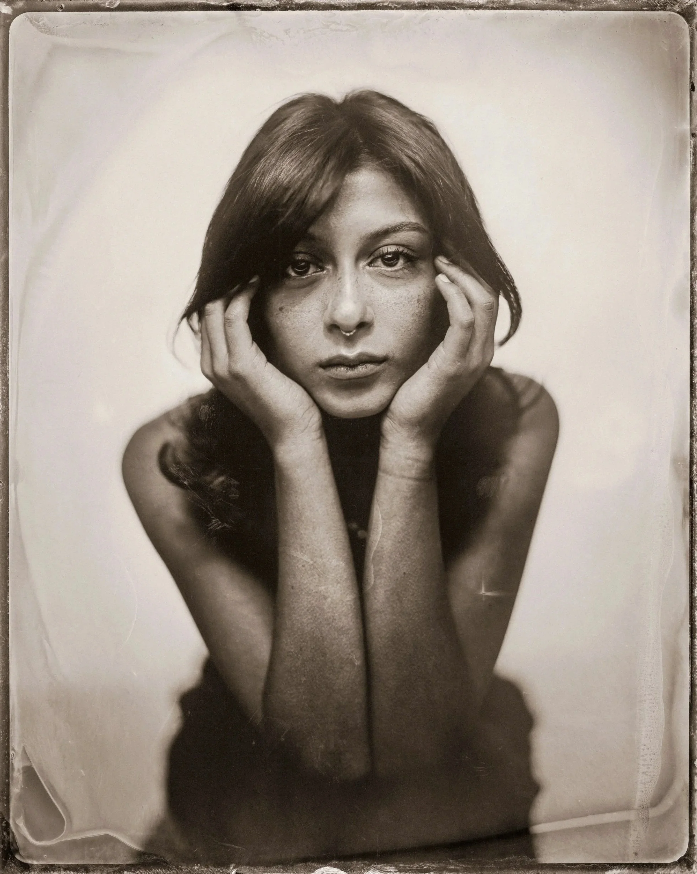 Black and white photograph of a young woman with long hair and freckles, resting her chin on her hands and looking directly at the camera.