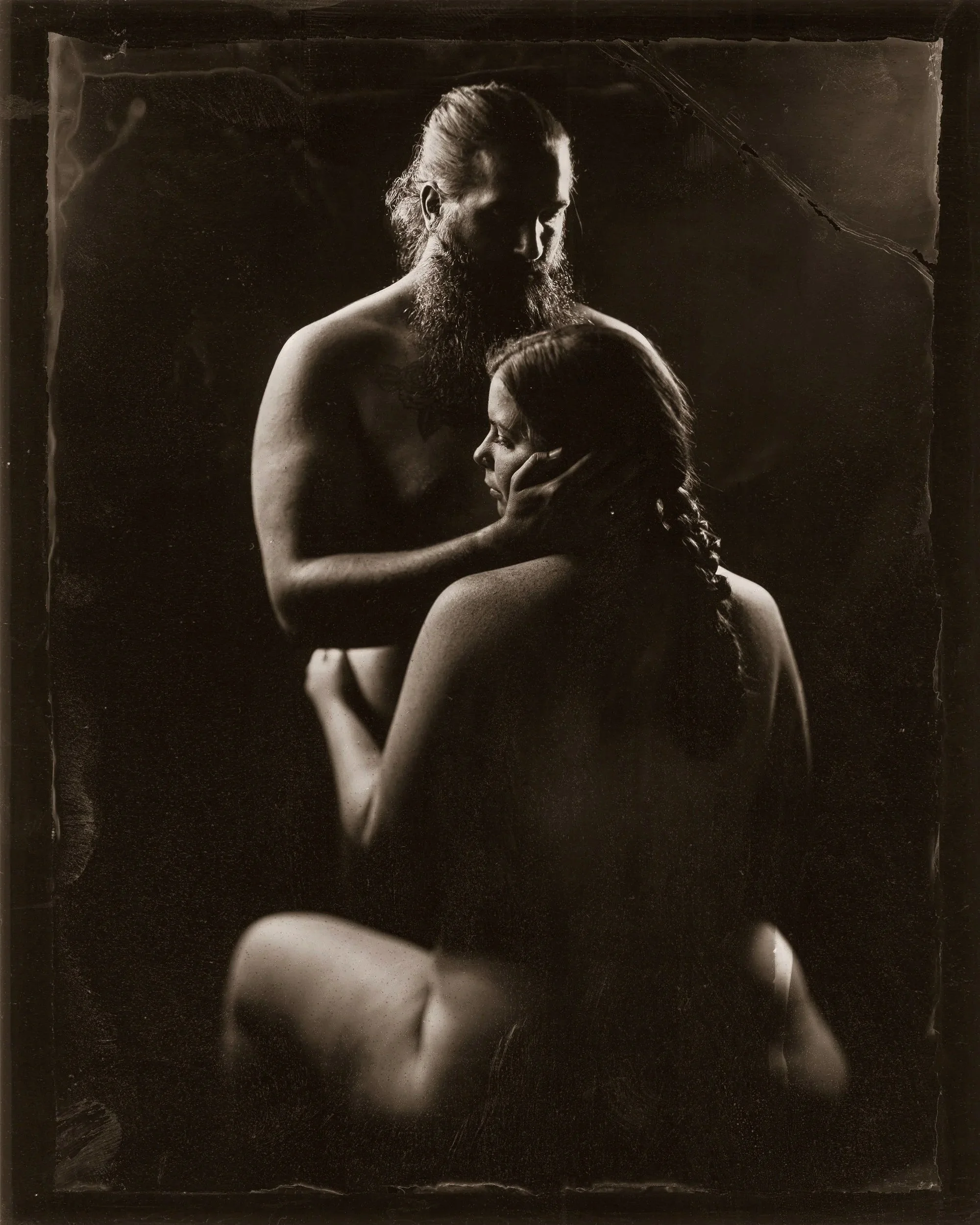 Black and white photograph of a shirtless man with a beard and long hair holding a woman with long hair, in an intimate pose. The woman's face is turned to the side and resting on the man's chest. The background is dark and plain.