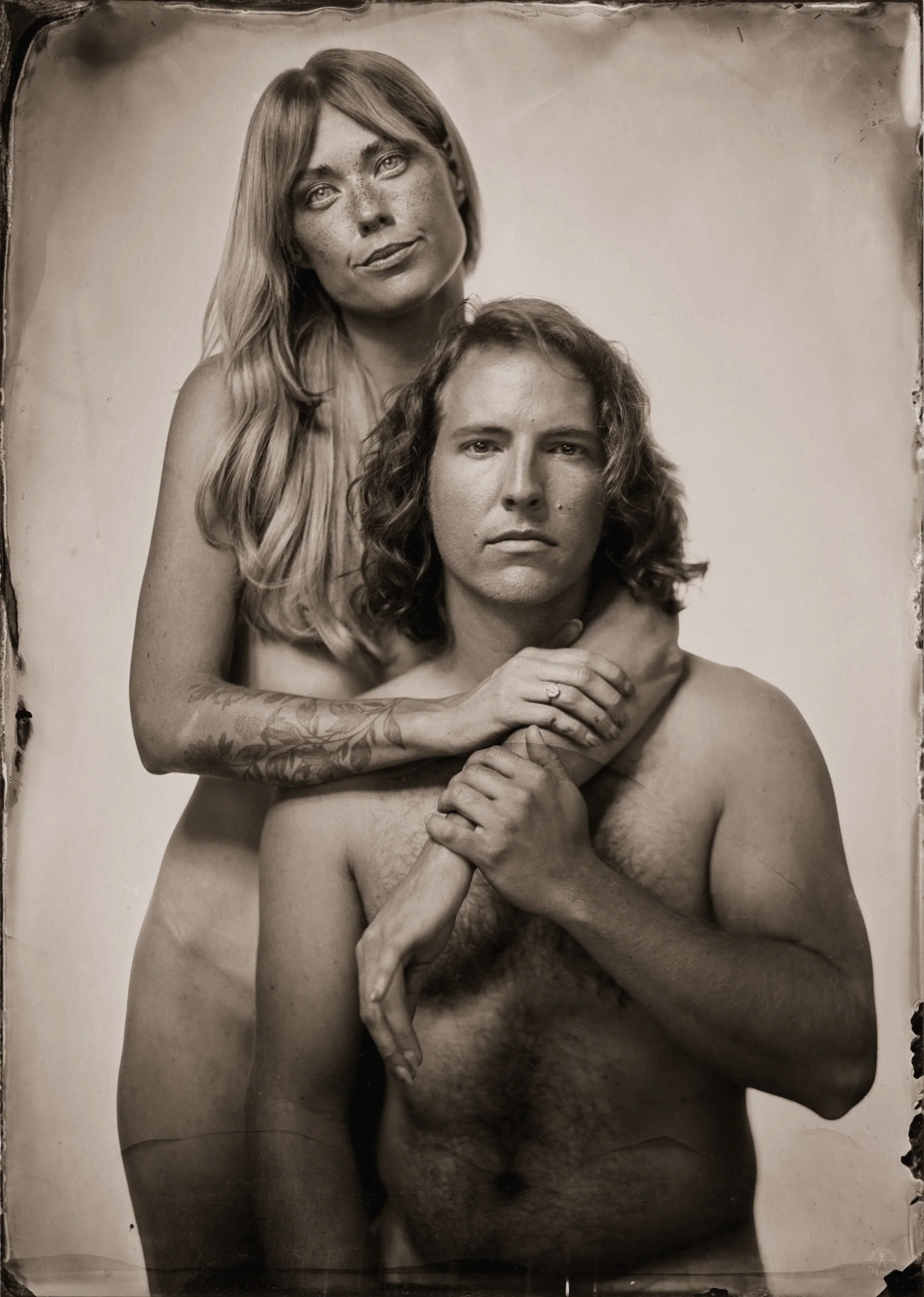 Black and white tintype photograph of a woman with long hair and freckles standing behind a shirtless man with long curly hair, with her arms around his neck, and their hands clasped in front of his chest.