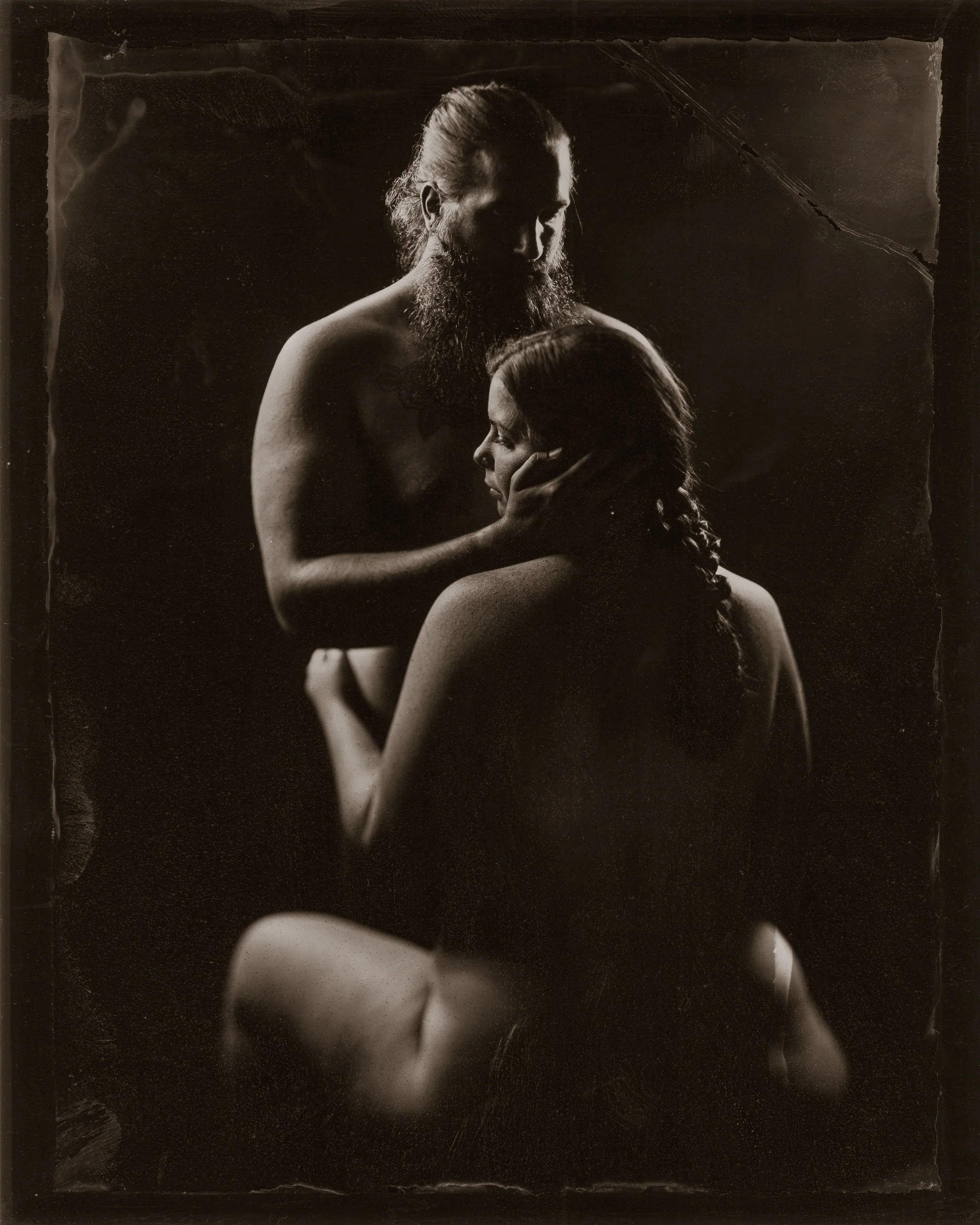 Black and white photo of a bearded man with long hair embracing a woman with long hair, both topless, in an intimate pose.