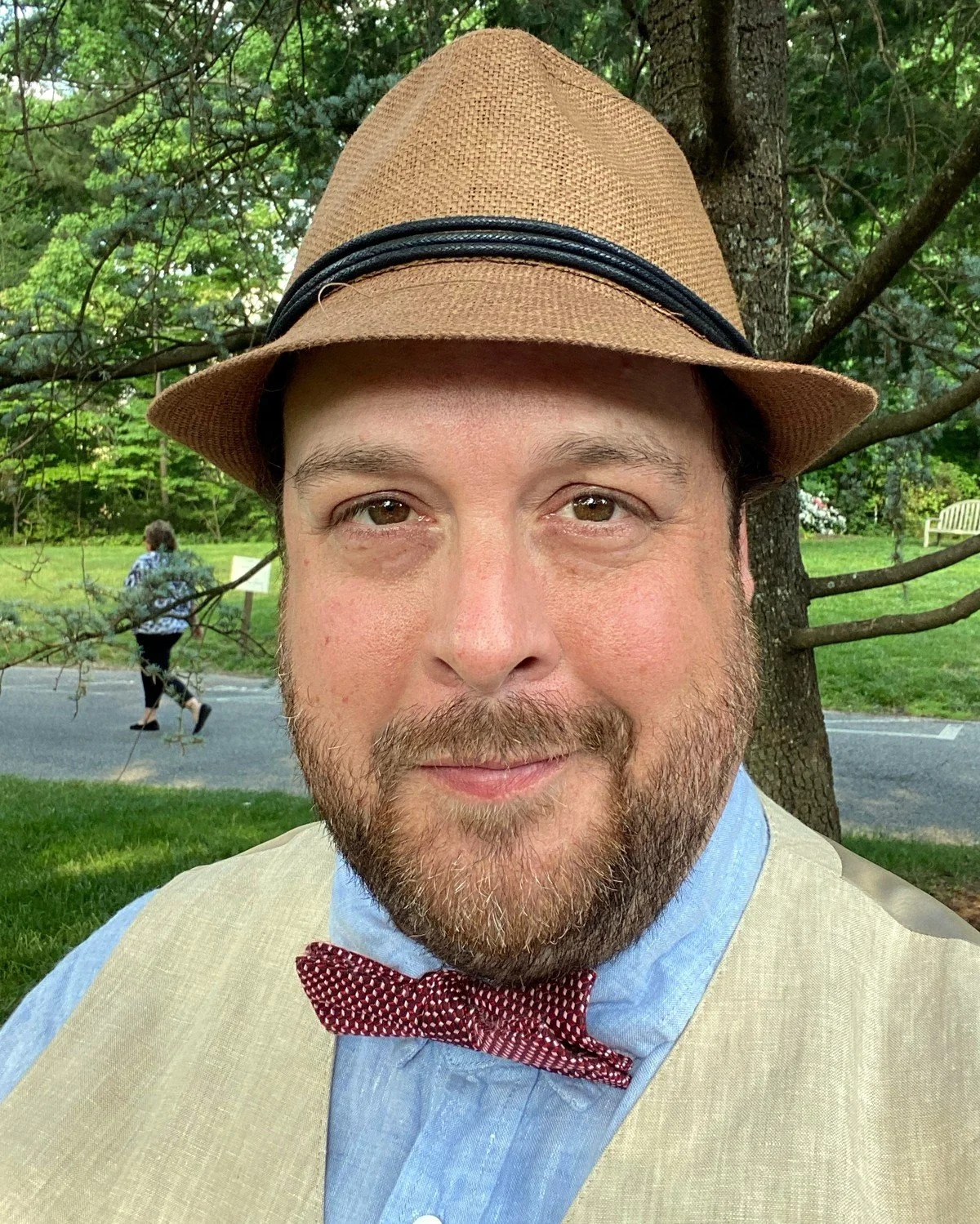 A man with a beard wearing a tan fedora hat, a beige vest, a light blue shirt, and a red patterned bow tie, smiling outdoors near a tree with green foliage, and a woman walking in the background on a sidewalk.