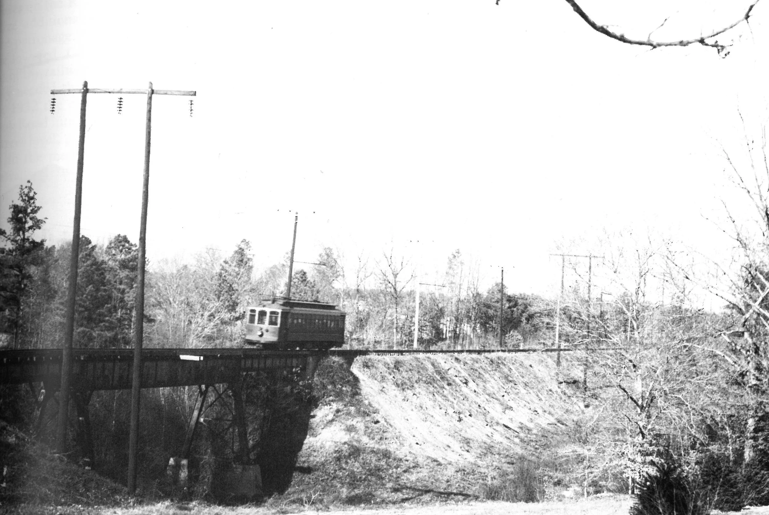 Black and white photograph of a train crossing a bridge over a forested area, with telephone poles and trees in the background.