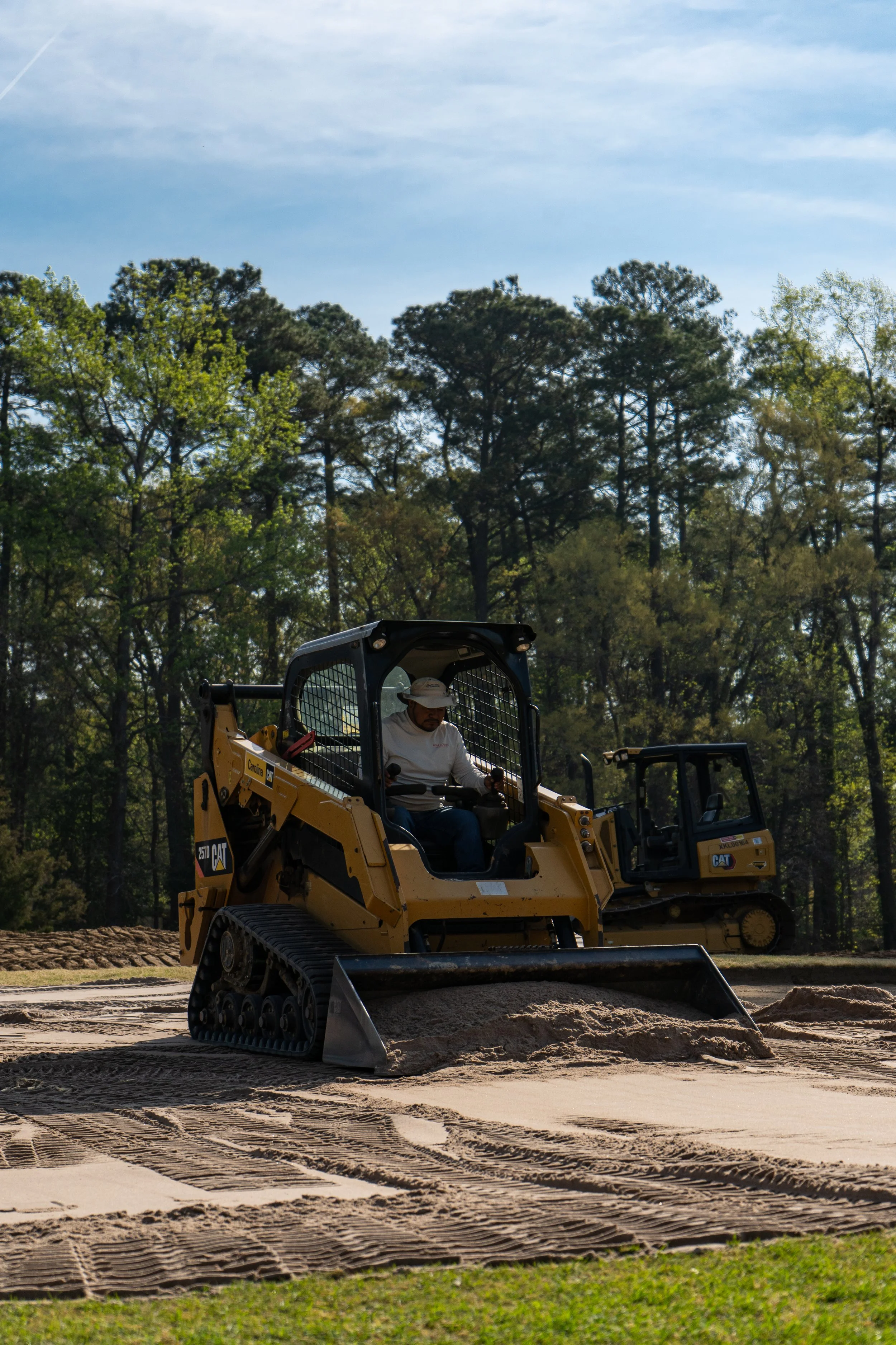 A construction worker operating a yellow Caterpillar bulldozer on a dirt construction site with trees and a blue sky in the background.