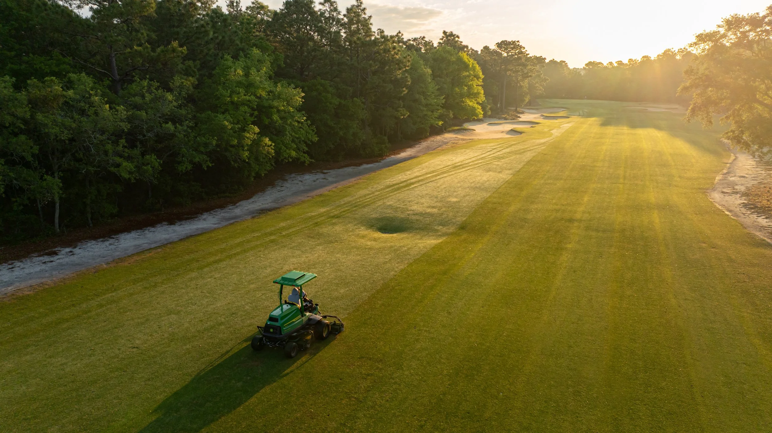 A golf course at sunset with a green lawn, sand traps, a water hazard on the left, trees in the background, and a small golf cart in the foreground. The sunlight creates long shadows across the grass.