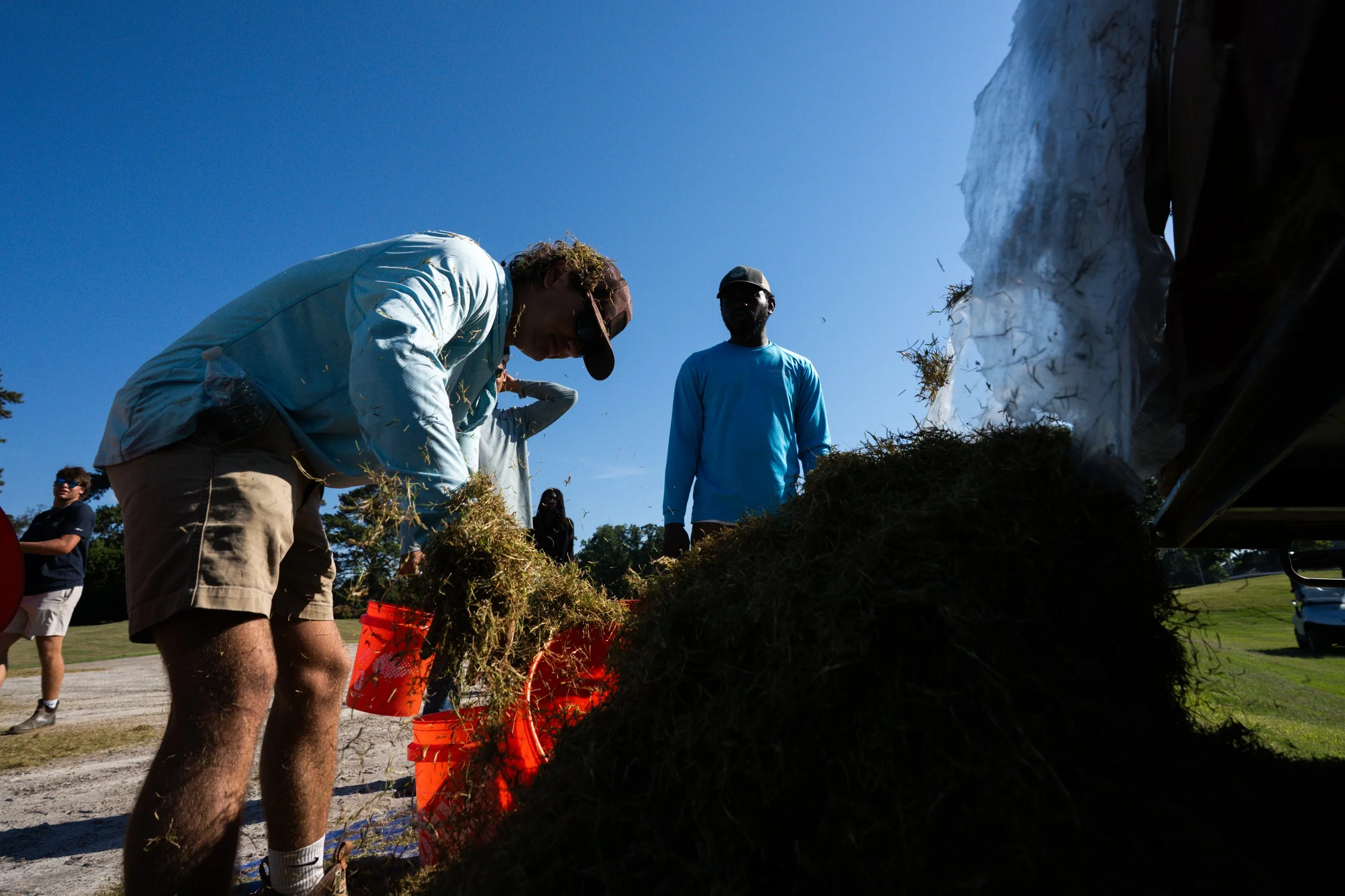 Group of people working outdoors, stacking or organizing hay or straw near a wall with a large white bag, on a sunny day.