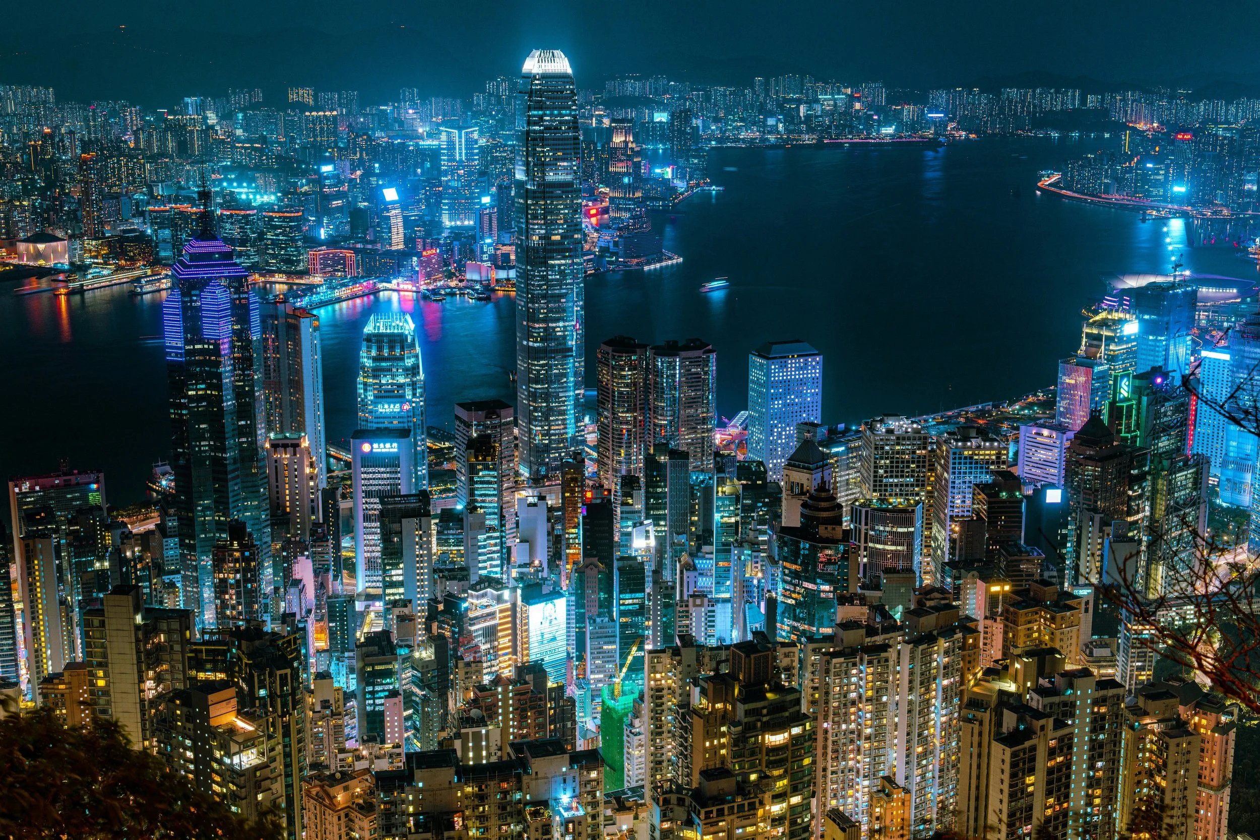 Night aerial view of a cityscape with illuminated skyscrapers and buildings along a river, featuring a prominent tall, rounded skyscraper, with brightly colored lights on the buildings and water reflections.