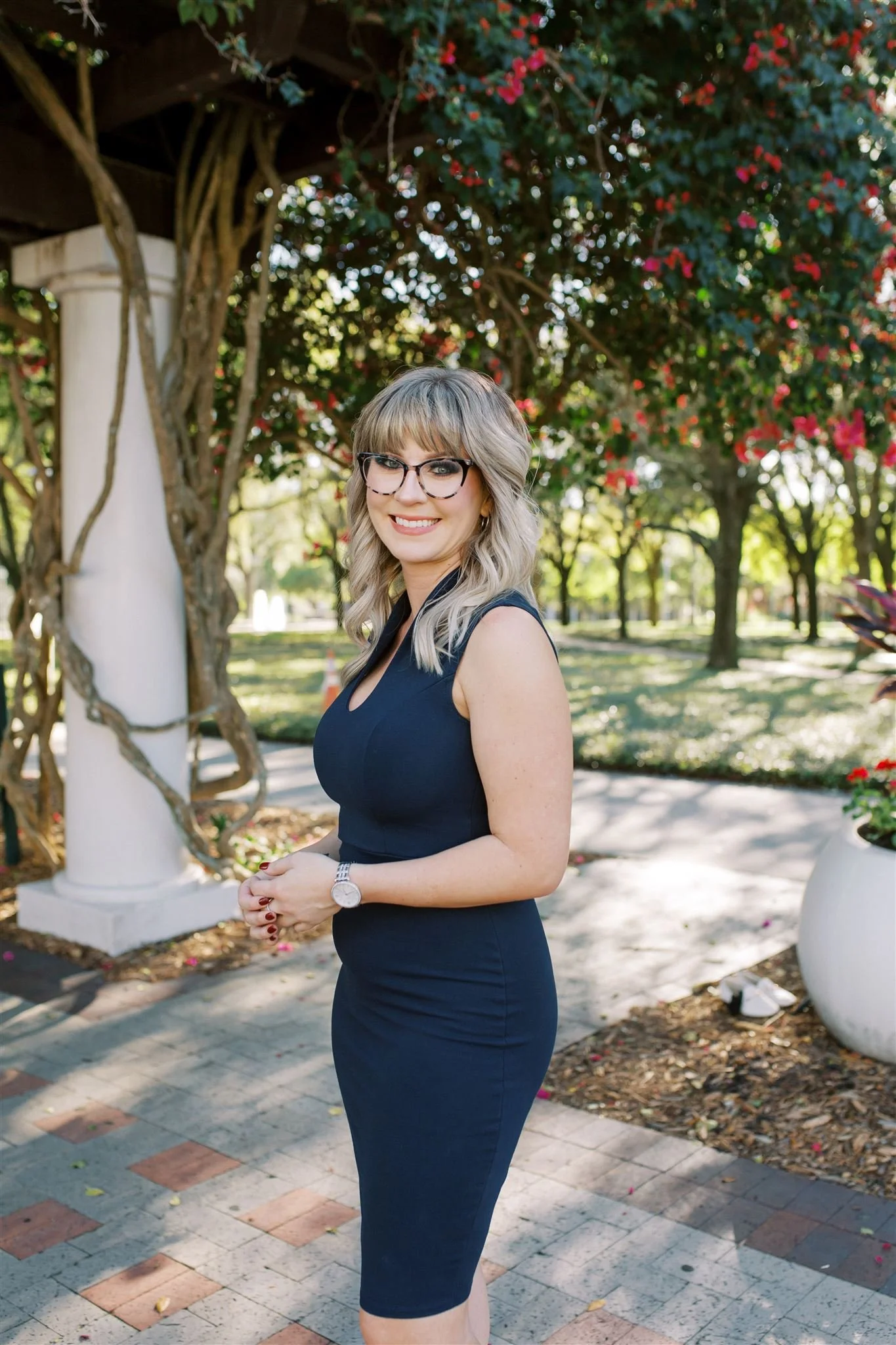 A woman with blonde hair, glasses, wearing a sleeveless navy blue dress, standing outdoors, smiling, with trees and pink flowers in the background.