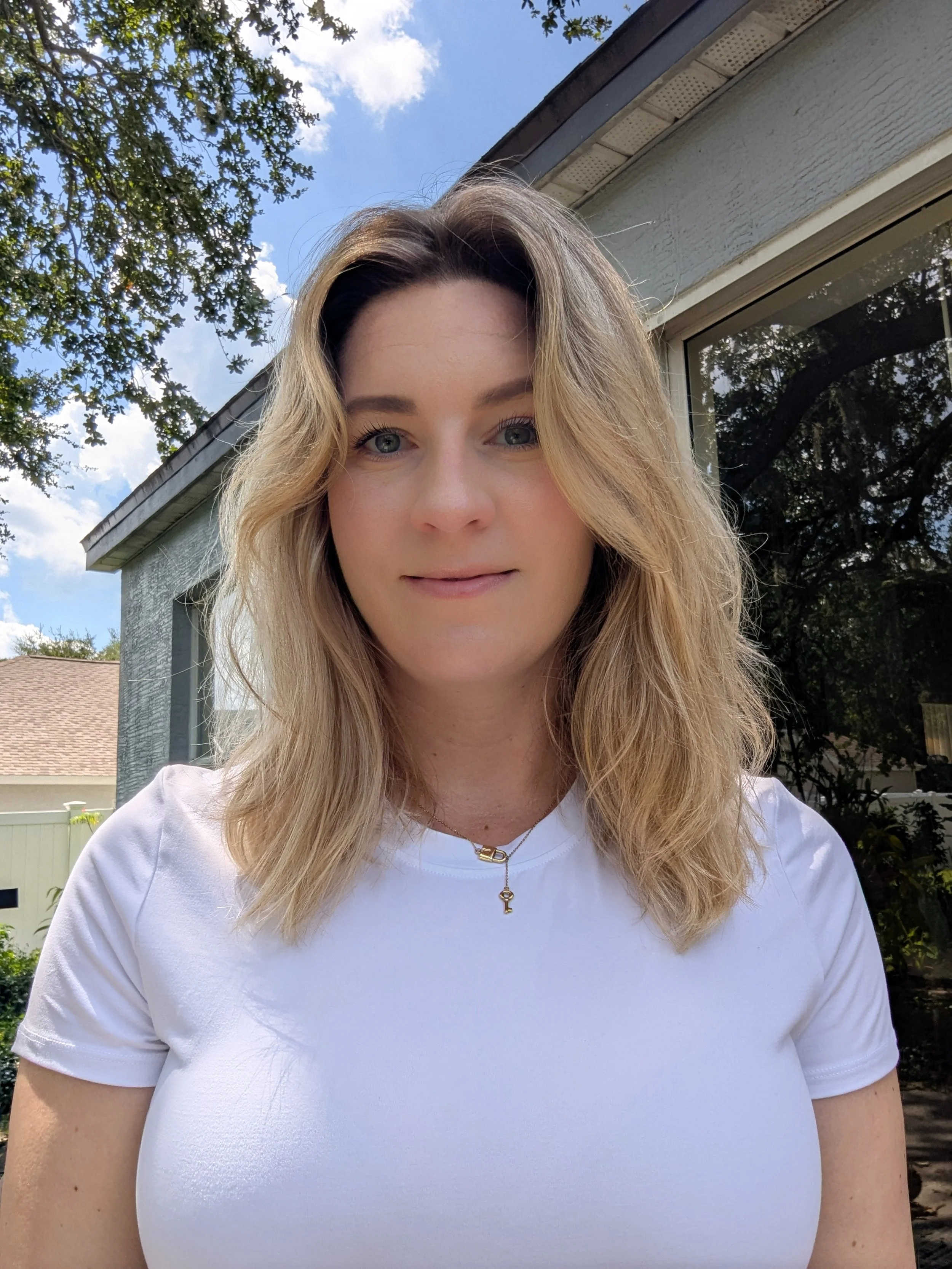 A woman with wavy blonde hair wearing a white shirt and a necklace with a key pendant standing outdoors on a sunny day.