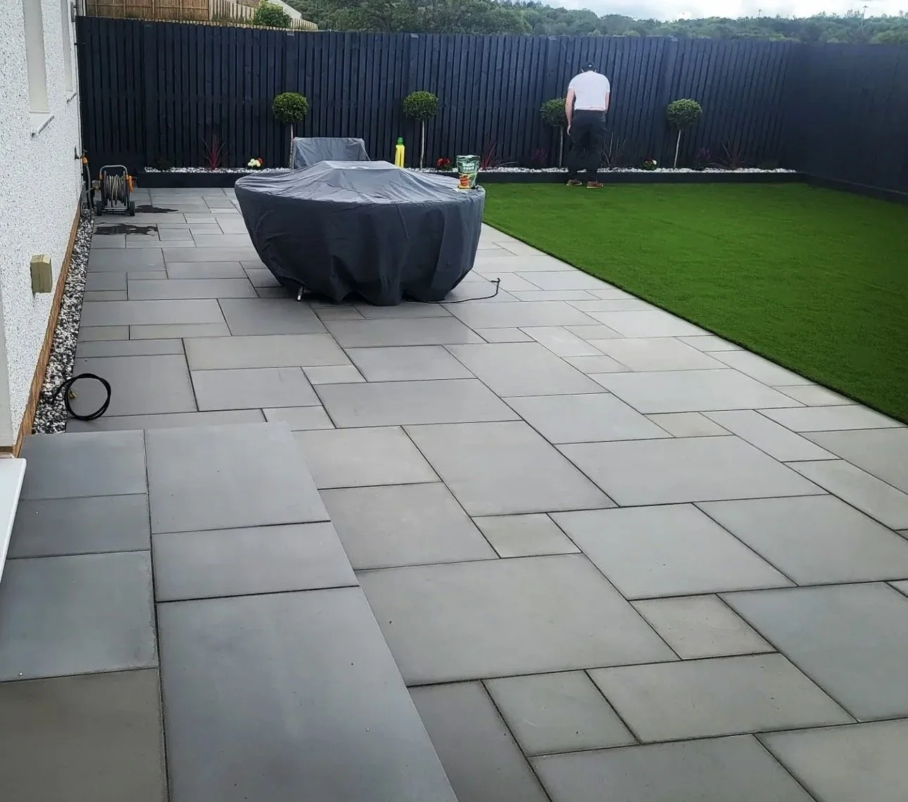 A backyard patio with large grey stone tiles, a grassy area, and a black fence. There's a covered round table with items on it, a person working near the fence, and some small decorative trees along the fence.